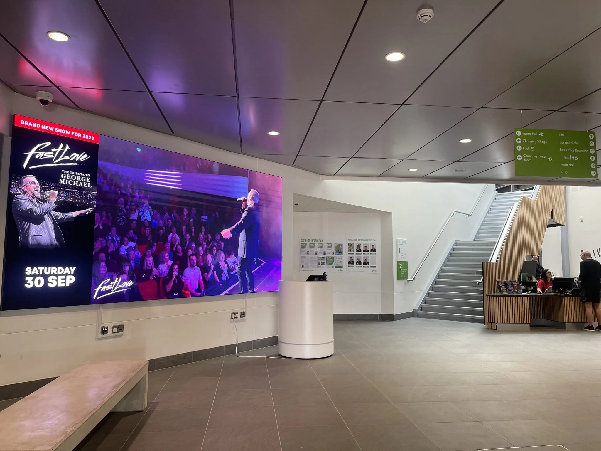 Hospital lobby with a large TV screen advertising a tribute concert for George Michael on Saturday, September 30, a reception desk with two staff members, a staircase leading upstairs, and green directional sign showing various facility locations.