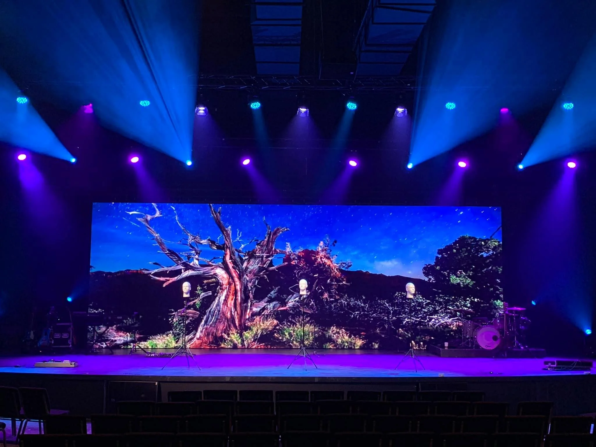 Stage with a large digital backdrop showcasing a desert landscape with a prominent, gnarled tree and a starry night sky. The stage is lit with blue and purple stage lights, and musical instruments such as microphones and drums are set up.