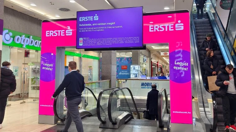 A shopping mall entrance with a bright pink archway advertising Erste Bank. People are using escalators and walking through the mall.