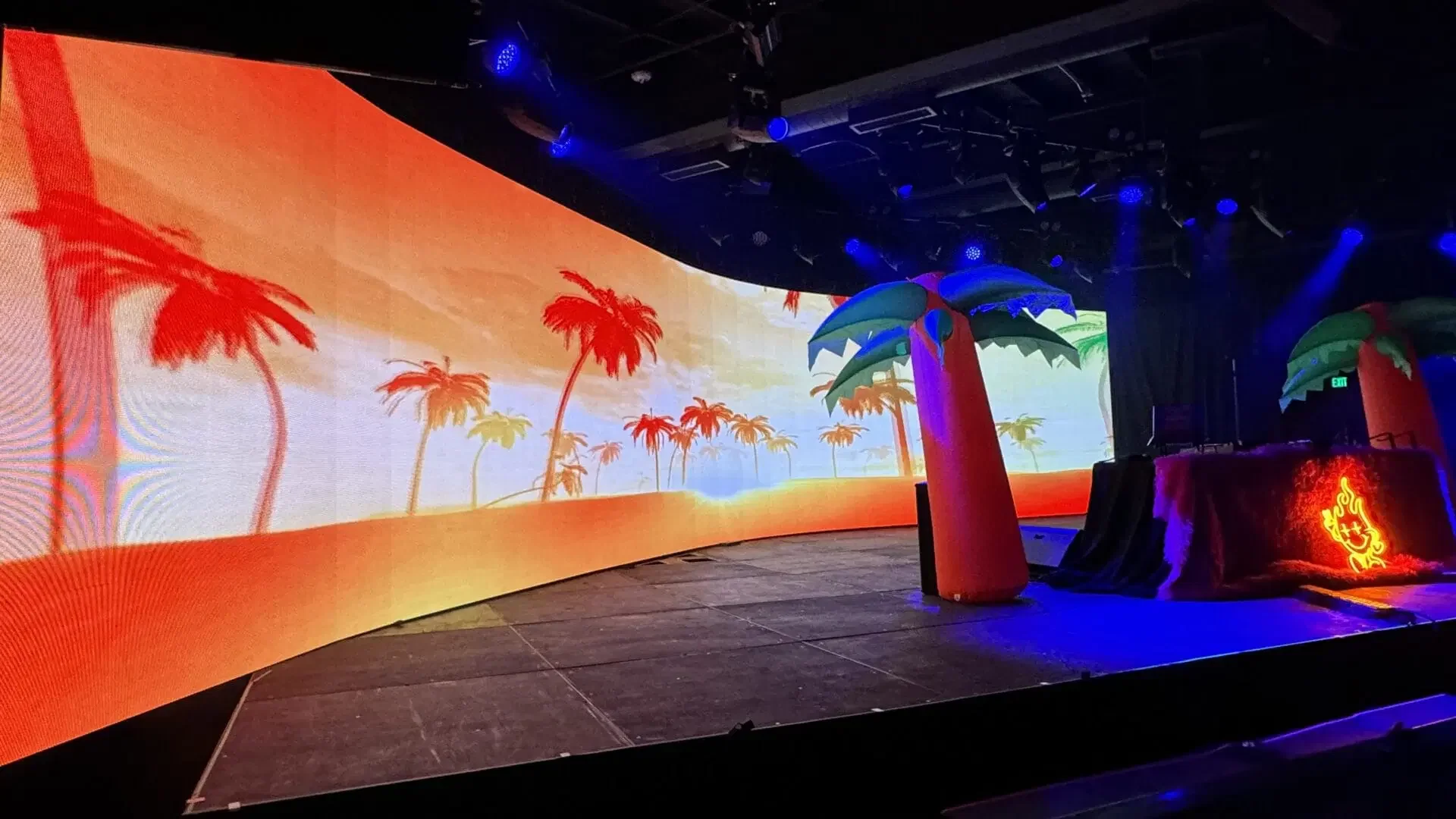 Stage setup with tropical theme, large LED screen displaying palm trees in sunset colors, inflatable palm trees, and DJ booth with neon graffiti logo, illuminated with blue and purple lighting.