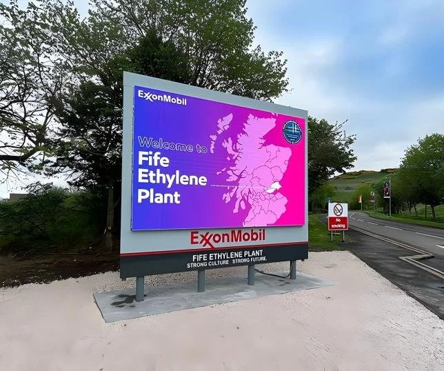 Large billboard welcoming visitors to the Fife Ethylene Plant, sponsored by ExxonMobil, with a map of Scotland and the plant's logo, situated near a road with trees in the background and a no smoking sign.
