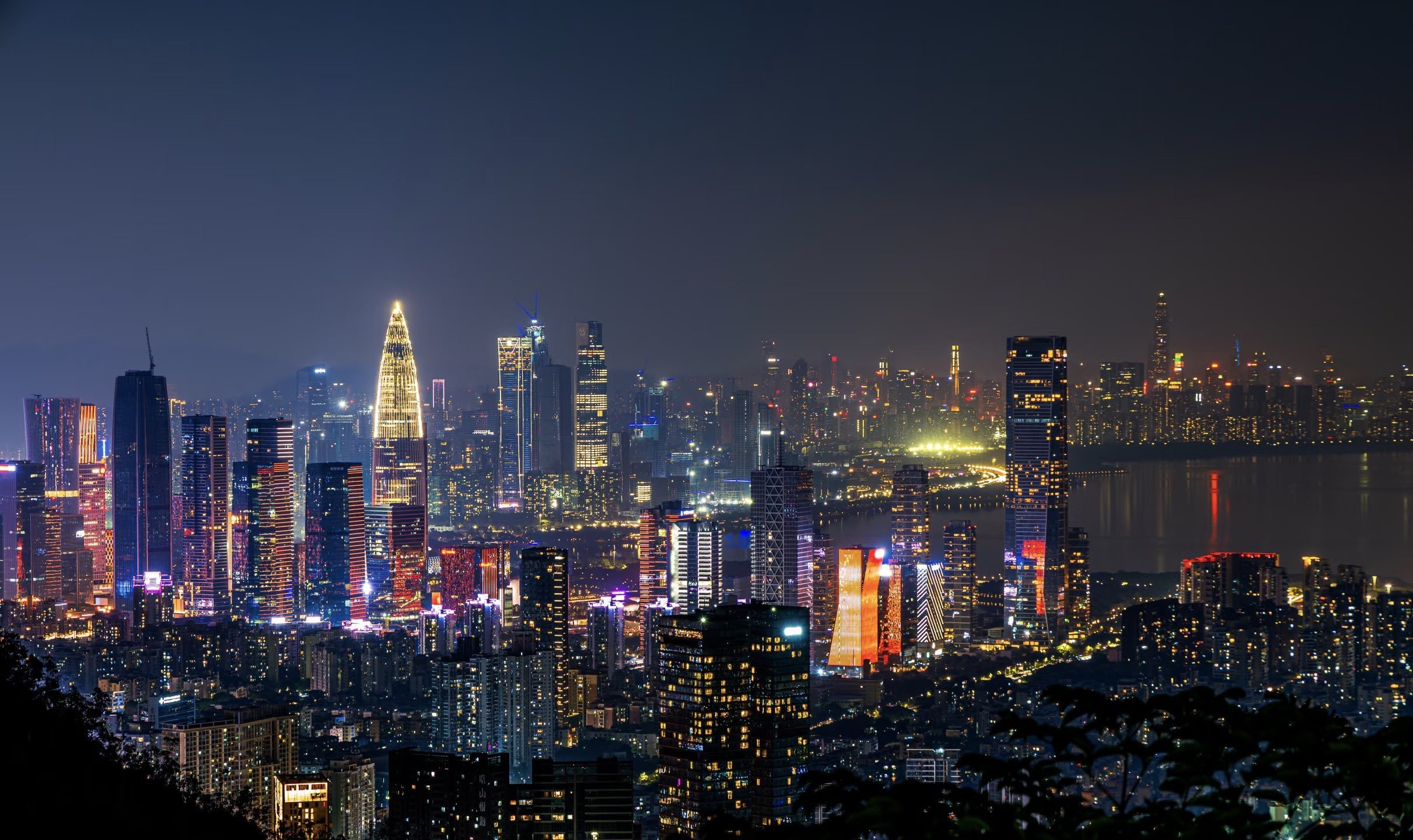 Nighttime city skyline with illuminated high-rise buildings and a body of water in the background.