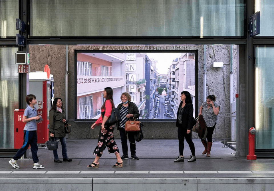 People standing and walking in an indoor corridor with a large screen showing an outdoor street scene in a city.