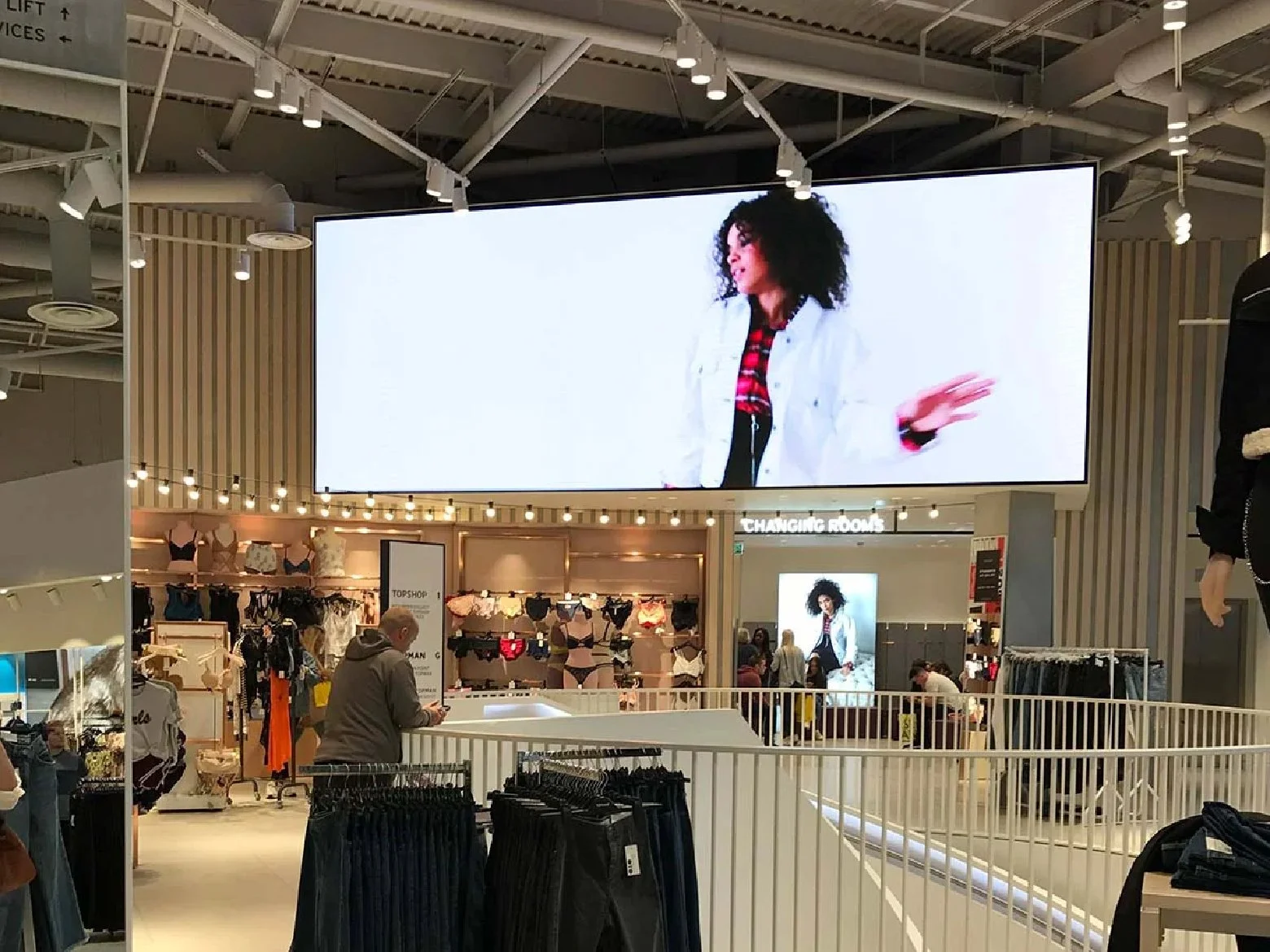 Large digital screen displays a woman with curly hair wearing a white jacket and red plaid shirt, gesturing with her right hand, in a retail clothing store.
