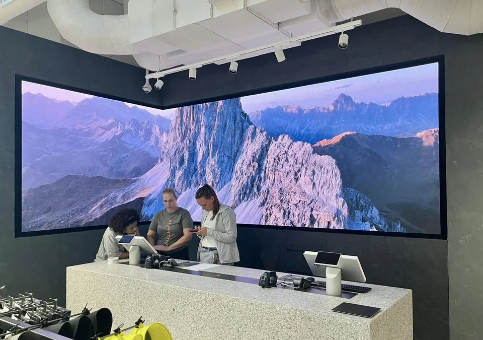 Three women are standing behind a counter in front of a large, curved digital display showing a mountain landscape.