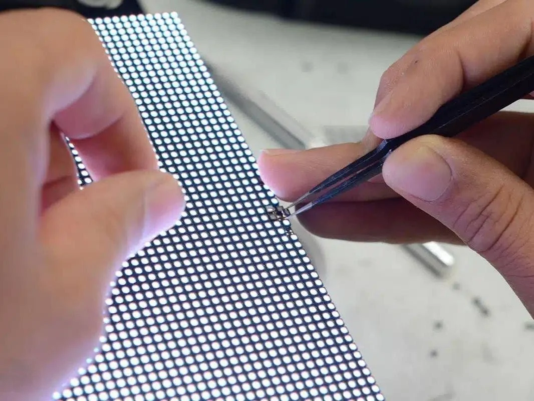 Close-up of a person using tweezers to repair or inspect a small electronic circuit on a perforated circuit board.