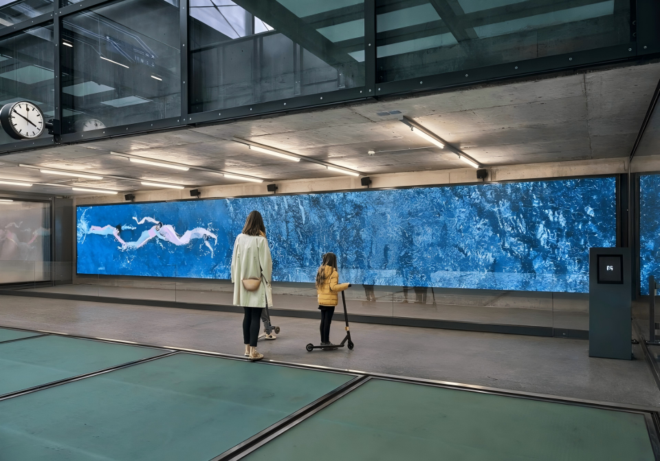An indoor public space with a large digital screen showing a swimming scene. A woman and a small girl with a scooter are observing the display. The area has modern architecture with glass and concrete, and a clock visible on the wall.