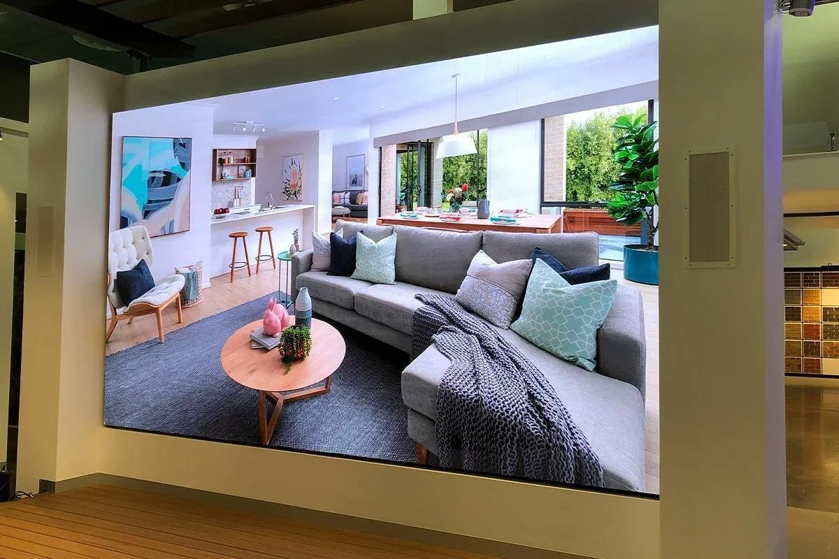 Living room with gray sofa, assorted pillows, and a round coffee table with decorative items, adjacent to an open kitchen and dining area with large windows and green outdoor views.