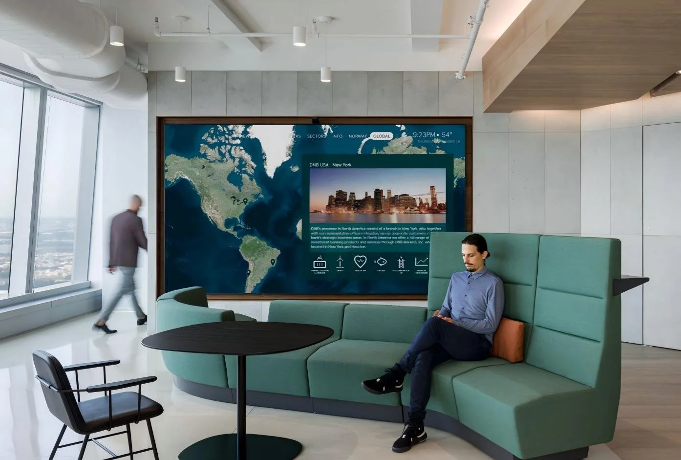 A man sitting on a green curved sofa in an office lounge area, looking at his phone. Behind him is a large digital display screen showing a world map and information about New York City. A person is walking past near large windows to the left.