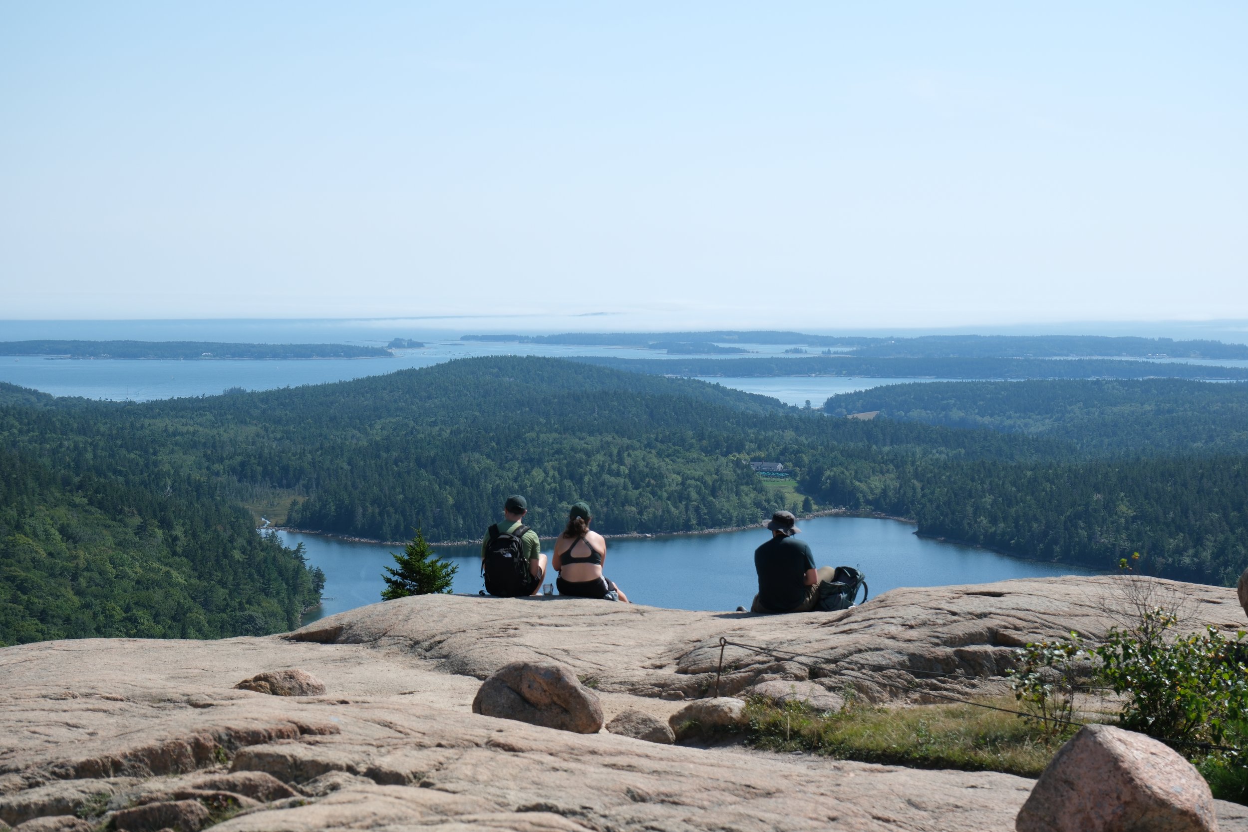 Three people sitting on a rocky ledge overlooking a scenic landscape of a forested area with a lake and water bodies in the distance on a clear day.