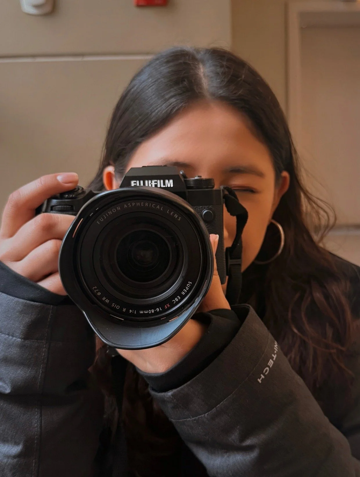 A woman taking a photo with a Fujifilm camera, partially covering her face and smiling, wearing a black jacket and hoop earrings.