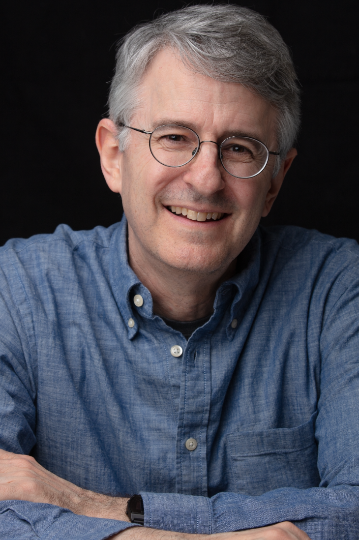 A smiling middle-aged man with salt-and-pepper hair and glasses, wearing a blue button-down shirt, sitting against a black background.