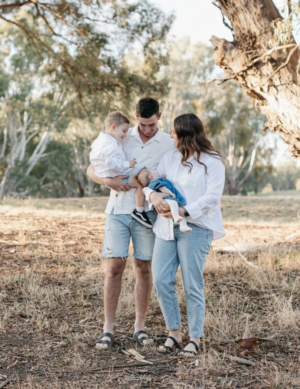 A family of four outdoors, with a man, woman, young boy, and baby, all dressed casually, standing on dry grass under a large tree.
