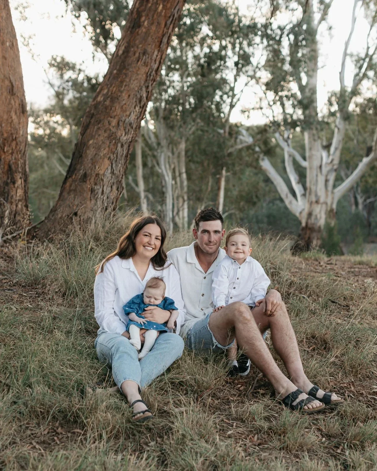 A family of four sitting on the grass in a wooded area during daytime. The mother and father are smiling, the mother holding a baby girl, and the older son sitting beside them.