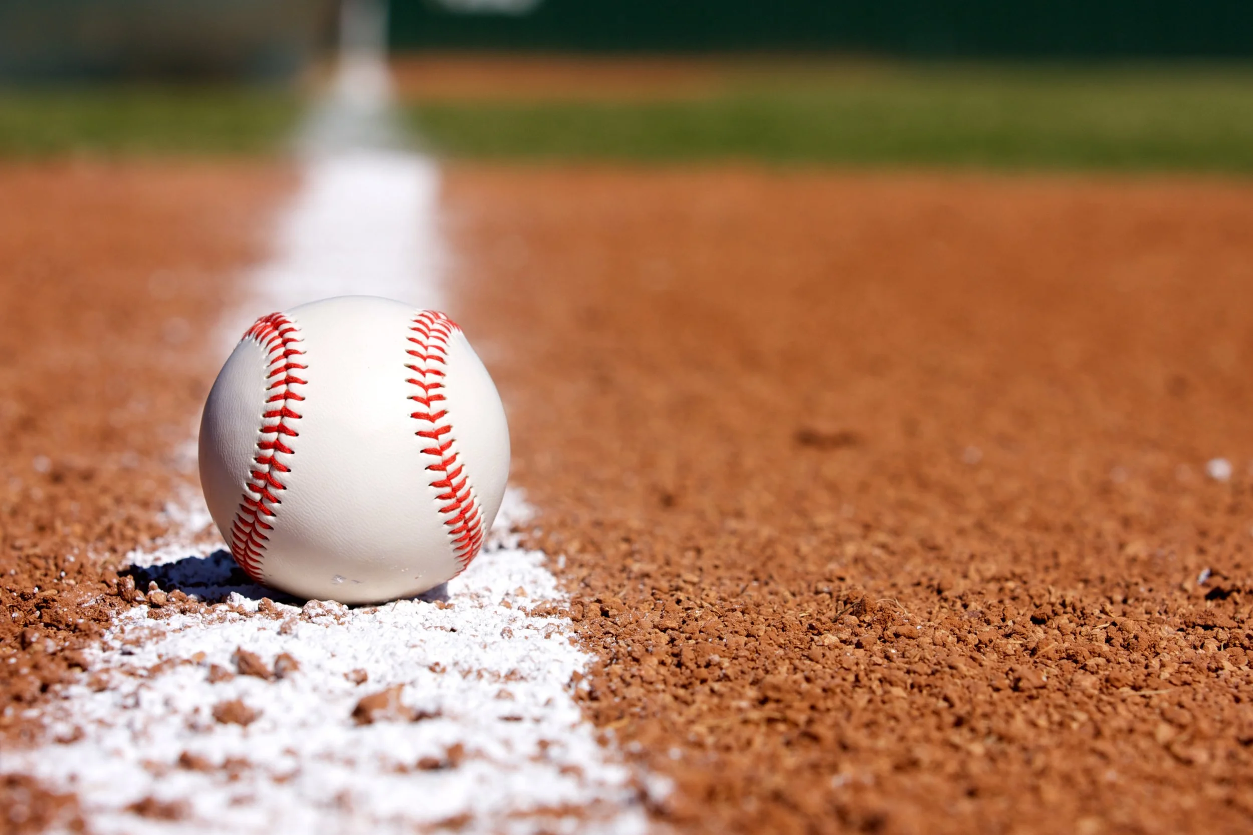 A close-up of a baseball resting on the white chalk line on a baseball field with the infield dirt surrounding it.