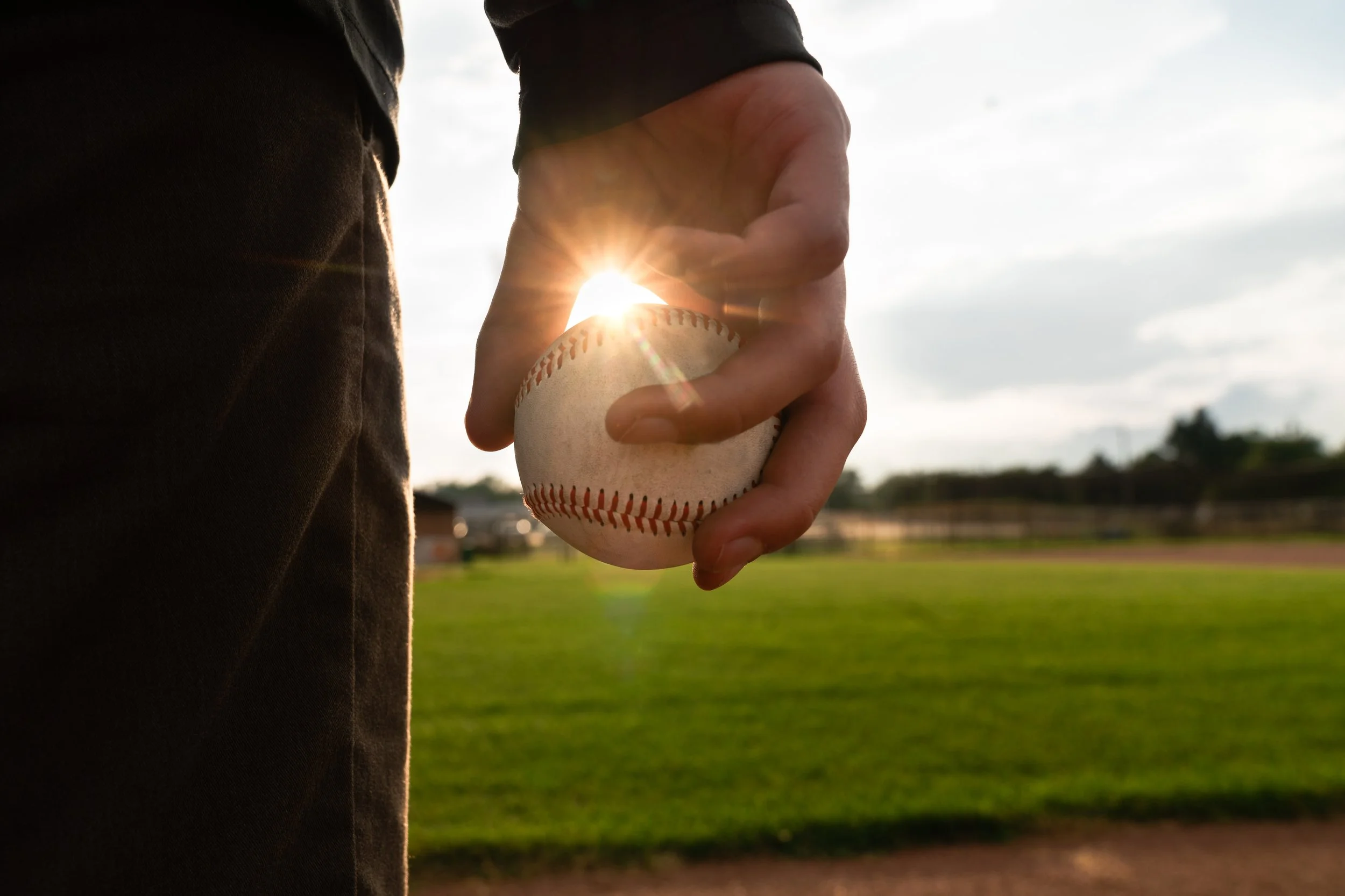 Close-up of a person holding a baseball in their right hand on a baseball field during sunset.