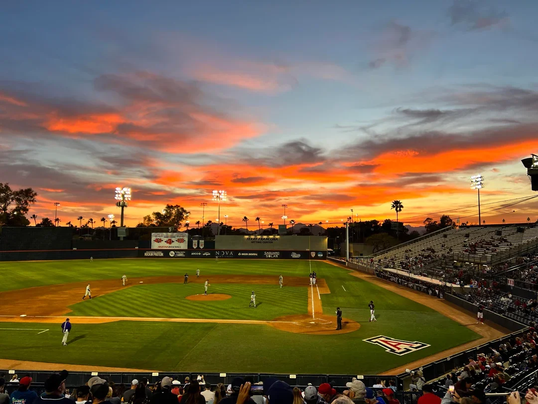 Baseball game at sunset with players on the field and fans in the stands.