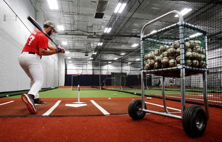 Person practicing baseball batting in an indoor training facility with a tee and baseballs, surrounded by netted cages and a cart filled with baseballs.
