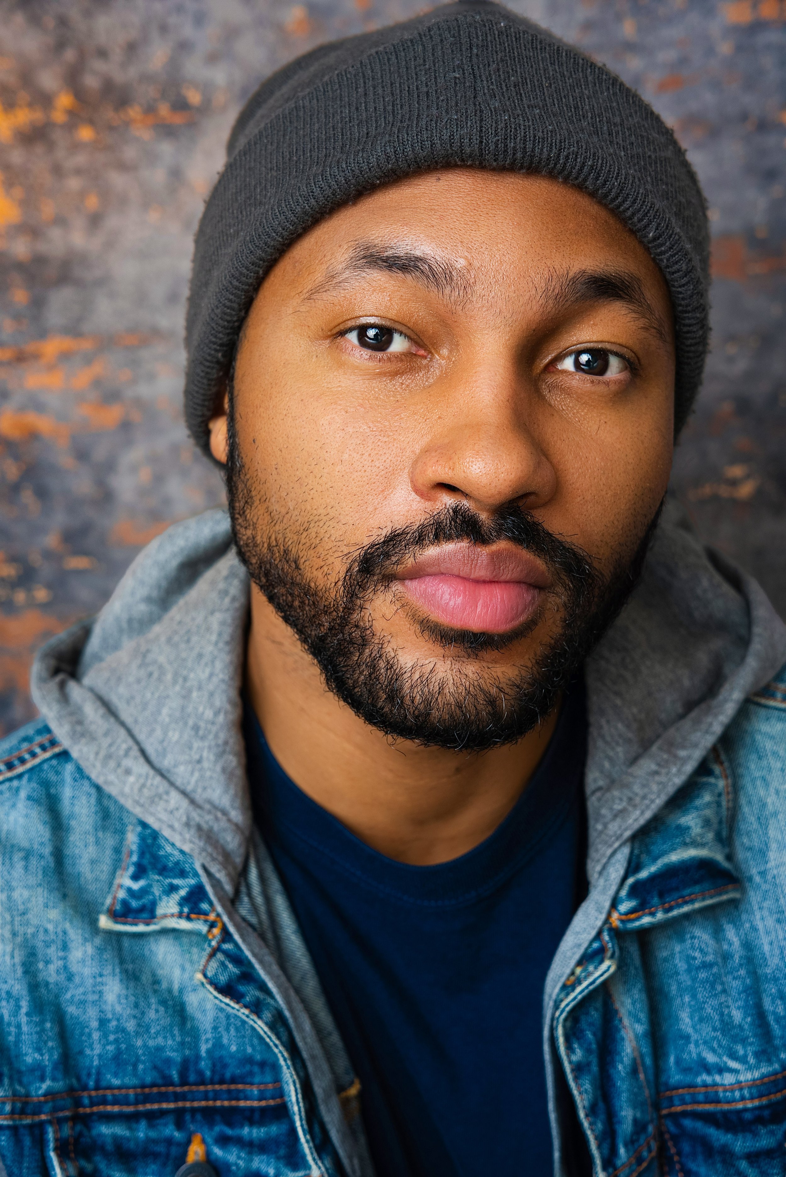 Close-up of a young man wearing a black beanie, denim jacket with gray hoodie underneath, and a navy T-shirt, against a dark, textured background.