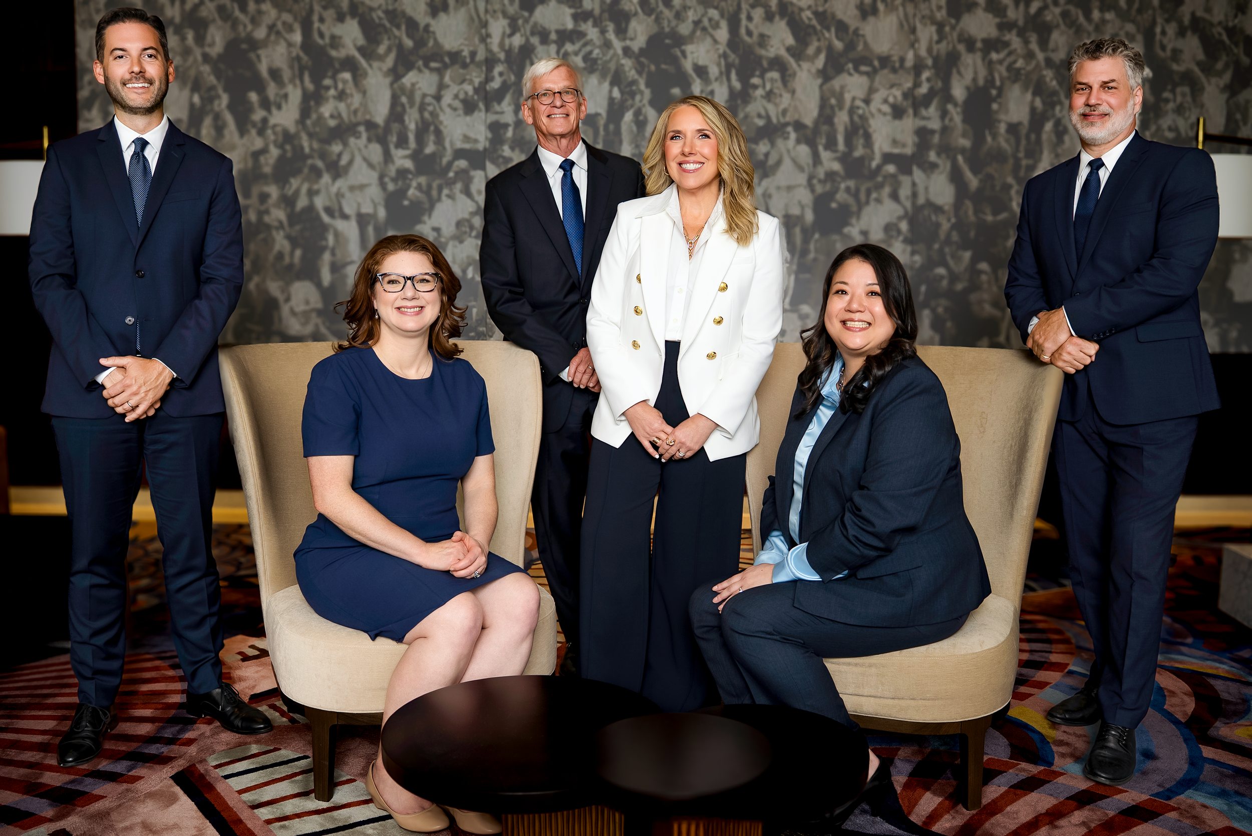 Group of seven professionally dressed people, four men and three women, posing in a hotel lobby or conference room.