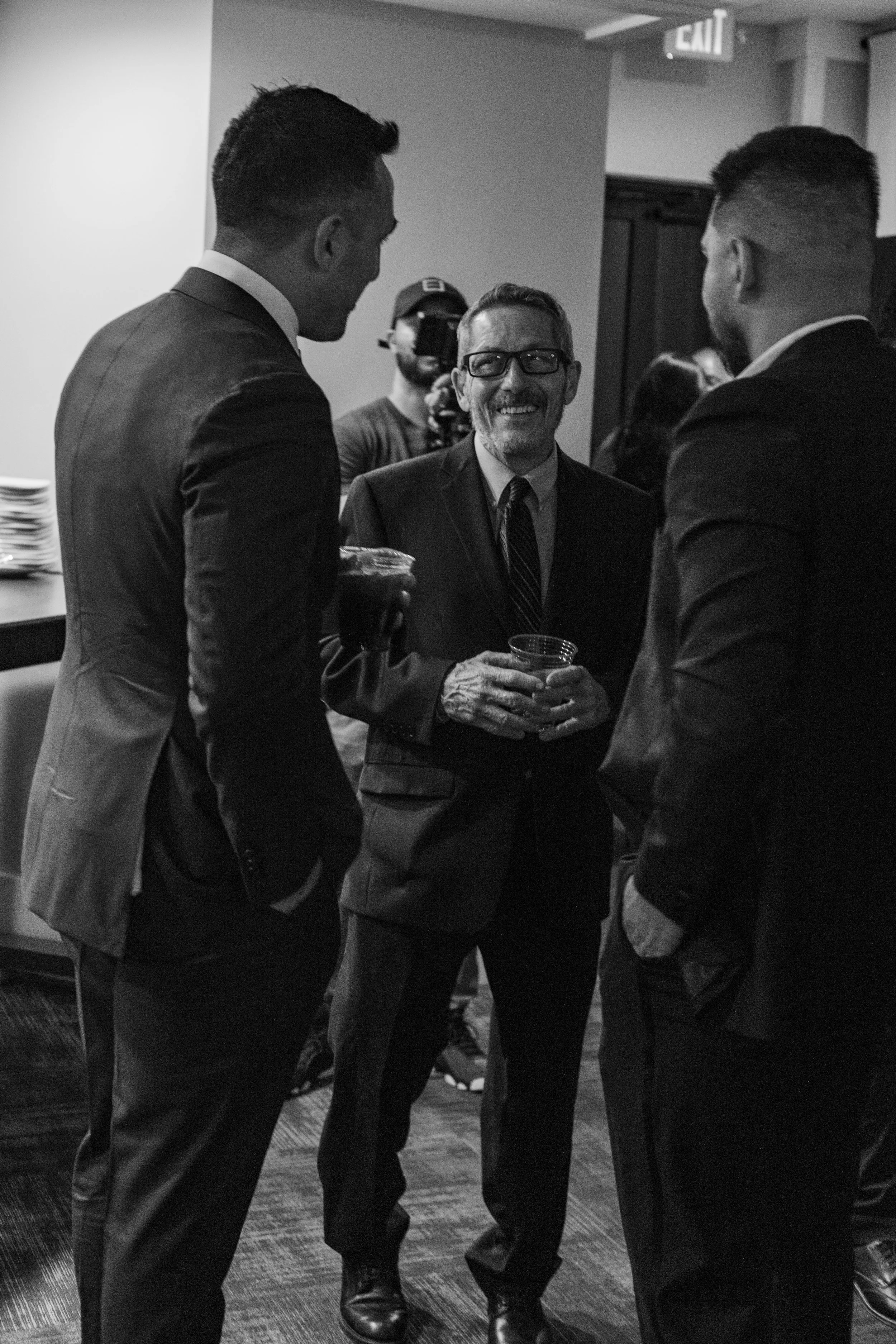 Three men in suits chatting at an indoor event, with a cameraman in the background.