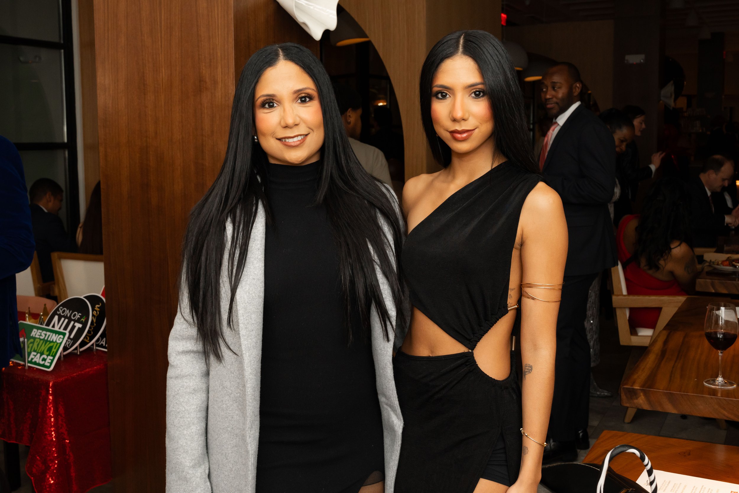 Two women posing together at an indoor event, one wearing a black turtleneck and gray coat, the other wearing a black one-shoulder dress with cutouts, with people in the background.