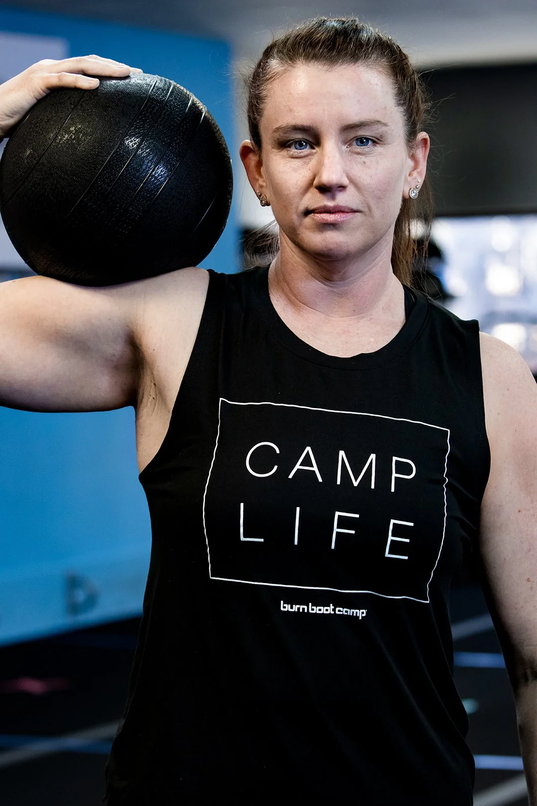 A woman holding a black medicine ball on her shoulder during a workout in a gym. She wears a black tank top with 'CAMP LIFE' written on it and has a serious expression.