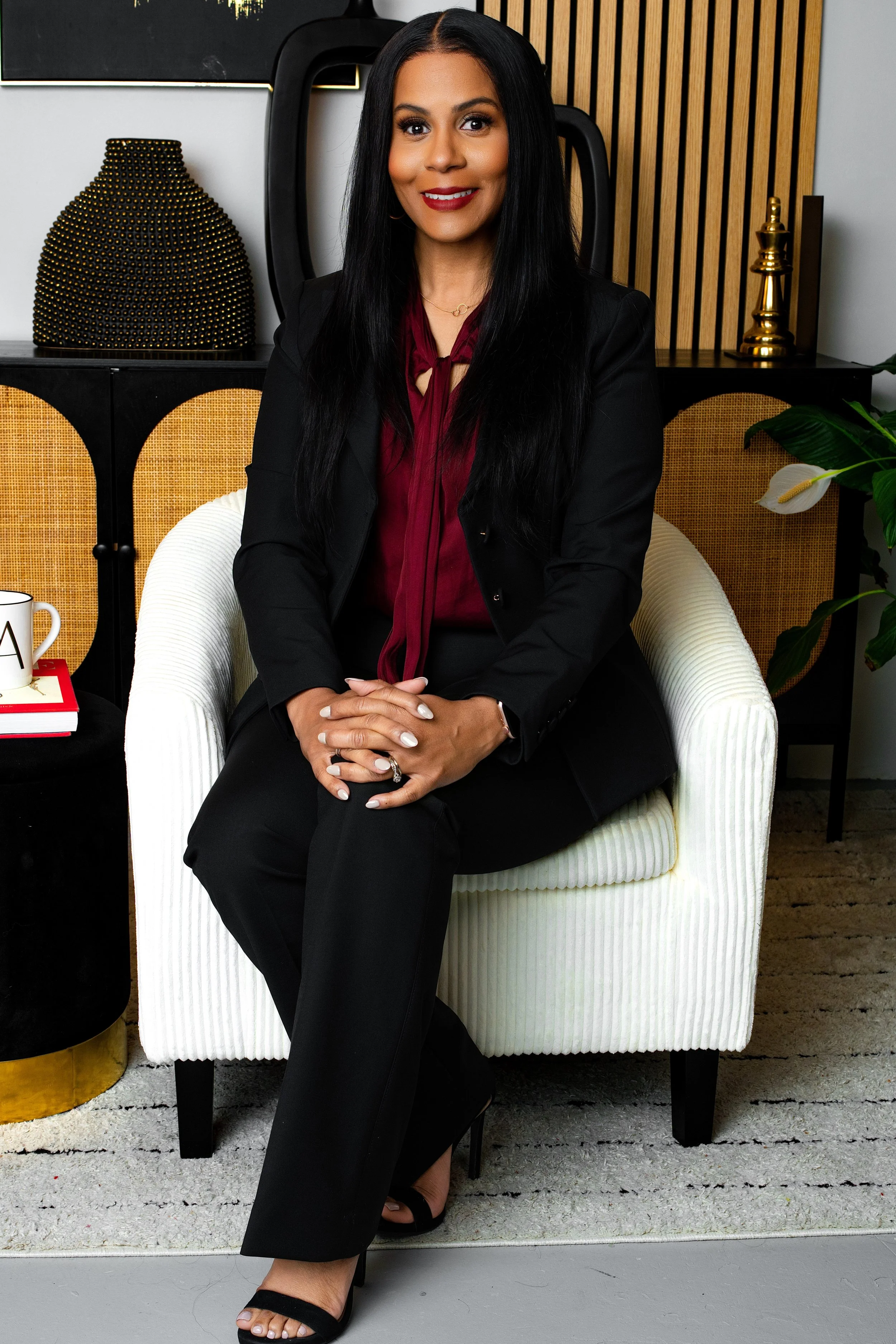 A woman with long black hair, wearing a black blazer, maroon blouse, and black pants, sitting on a white striped armchair in a modern office or living space with decorative items and plants.