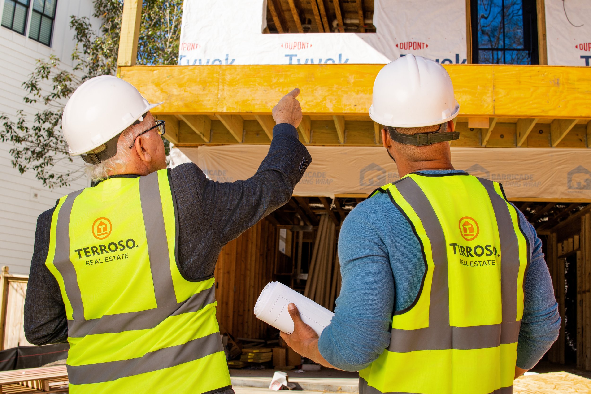Two construction workers on site, both wearing yellow safety vests and white hard hats, discussing plans while pointing at the second story of a house under construction.