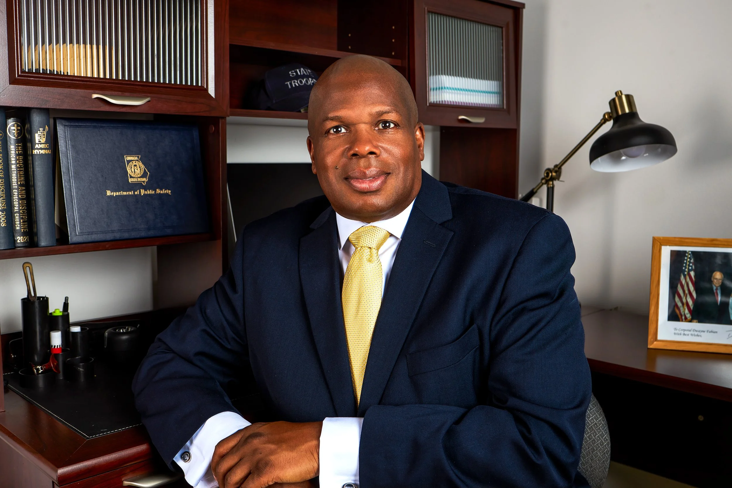 A man wearing a navy suit and yellow tie sitting at a desk in an office with books, framed photo, and a desk lamp.