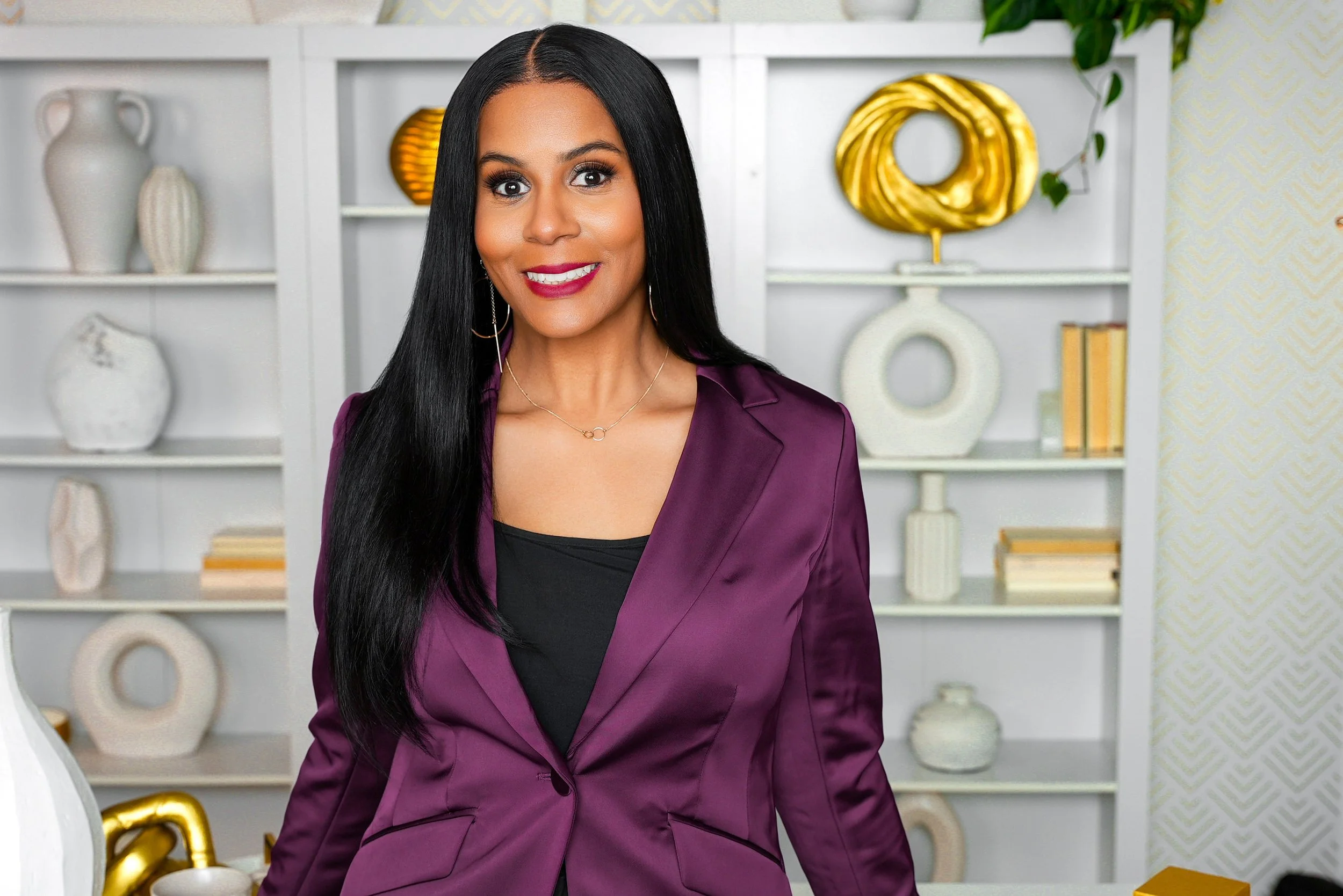 A woman with long black hair and a purple blazer smiling in a modern, decorated room with white shelves and decorative vases.