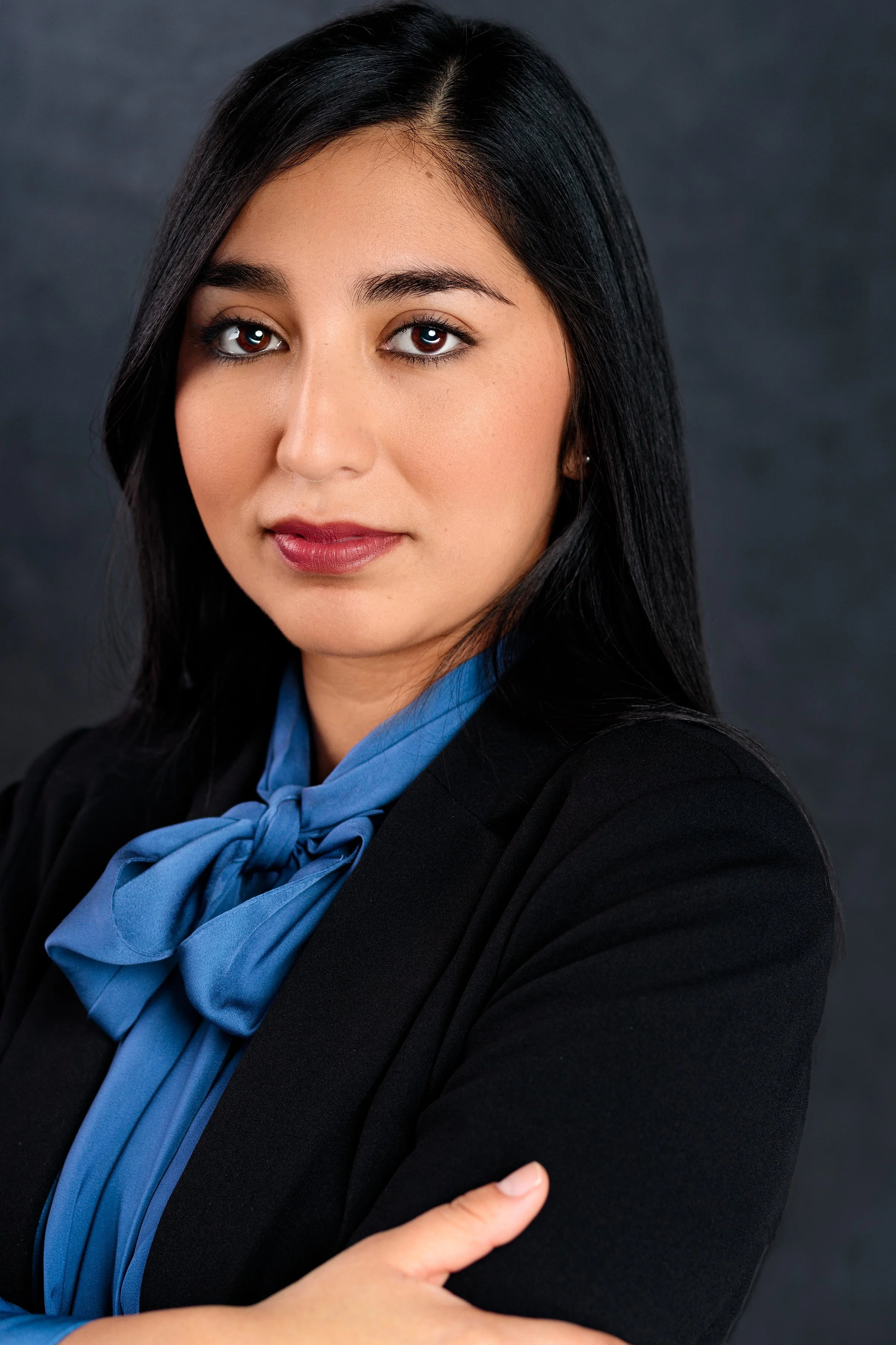 Professional woman with dark hair, wearing a black blazer and a blue scarf, posed with her arms crossed against a dark background.