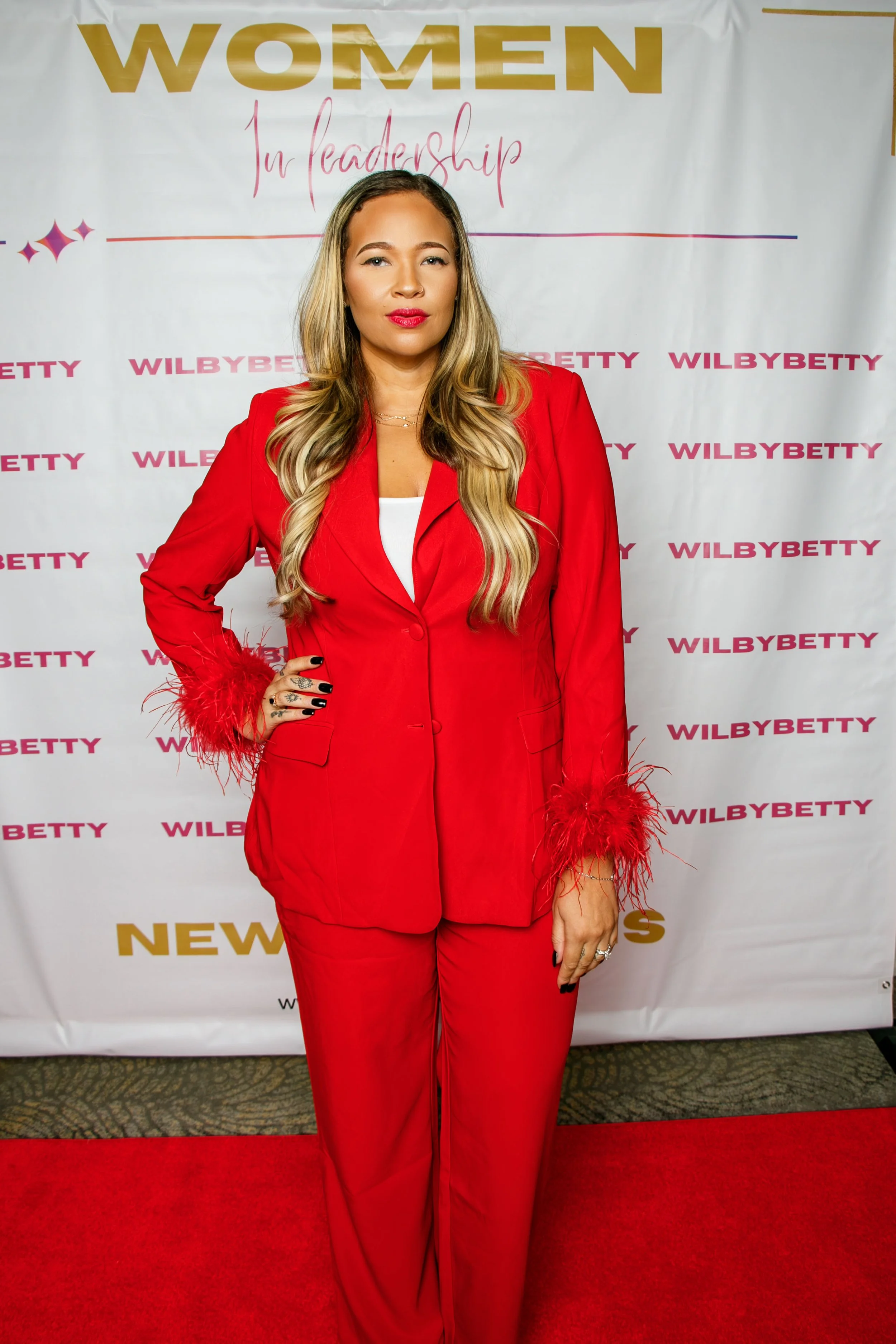 A woman with long blonde hair wearing a red suit with feathered cuffs, standing on a red carpet in front of a backdrop that says 'Women in Leadership' and 'WilbyBetty'.