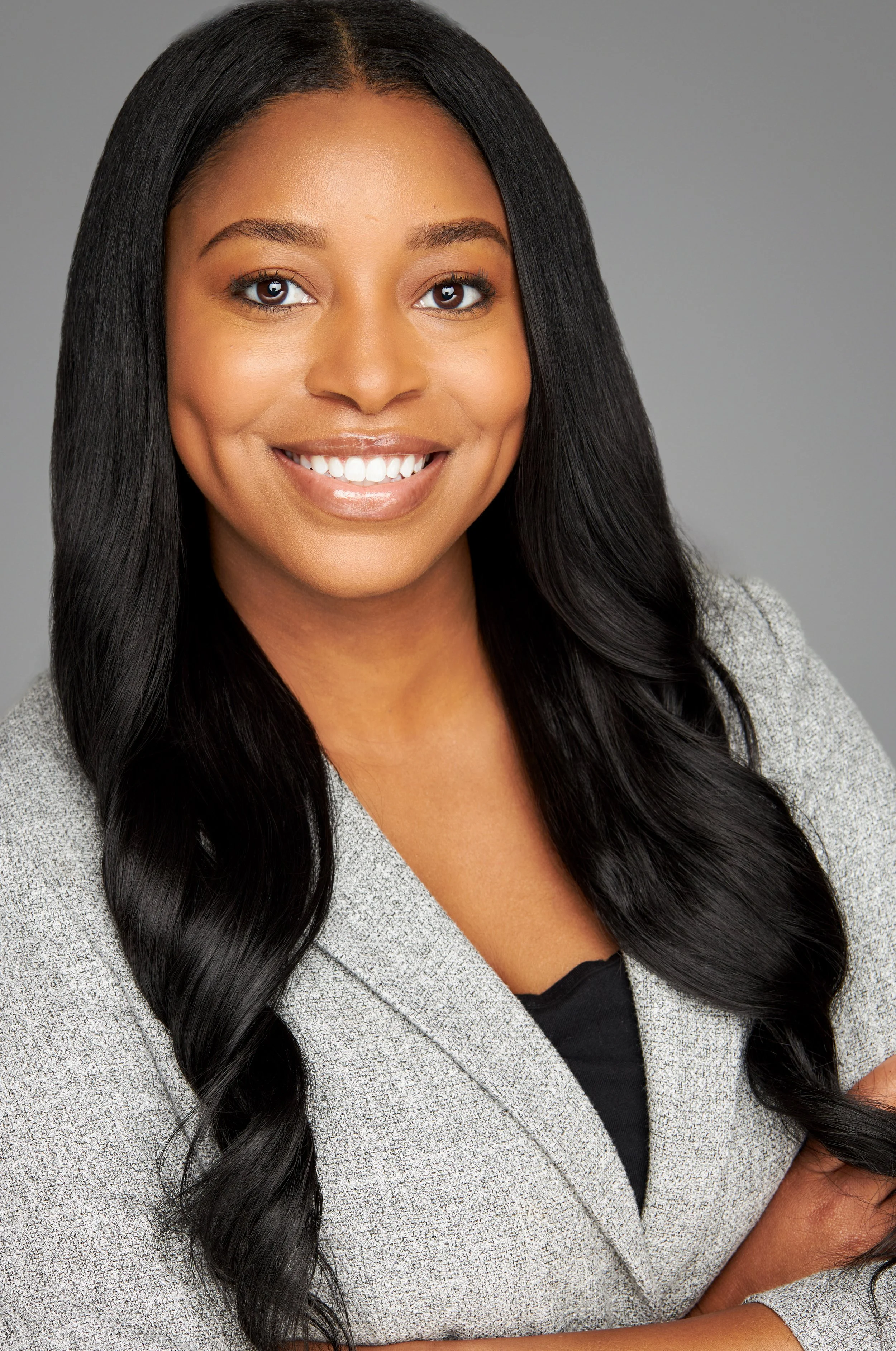 A smiling woman with long black hair wearing a gray blazer, posing against a plain gray background.