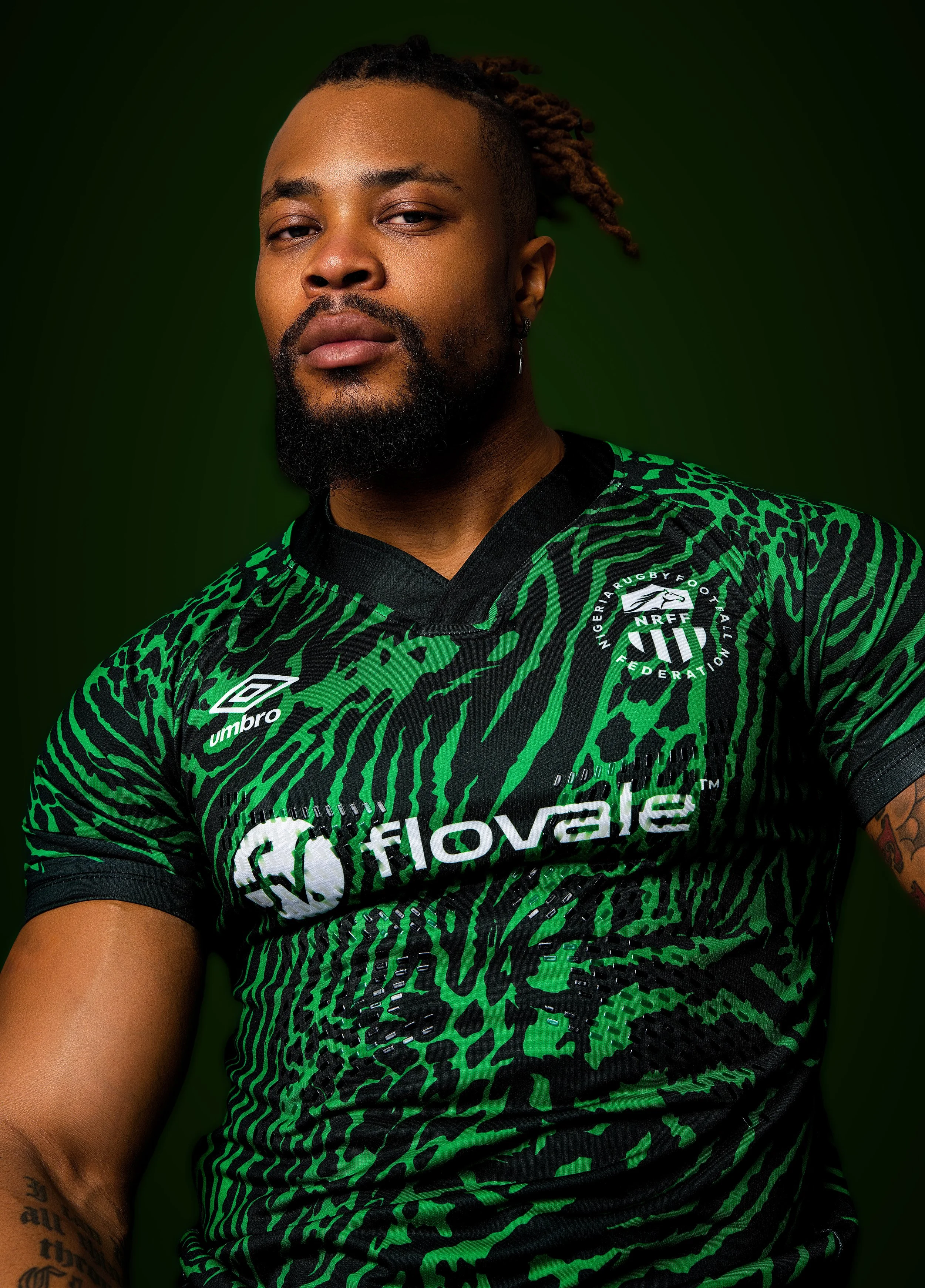 Portrait of a man with a beard and dreadlocks wearing a green and black rugby jersey with logos, against a dark green background.