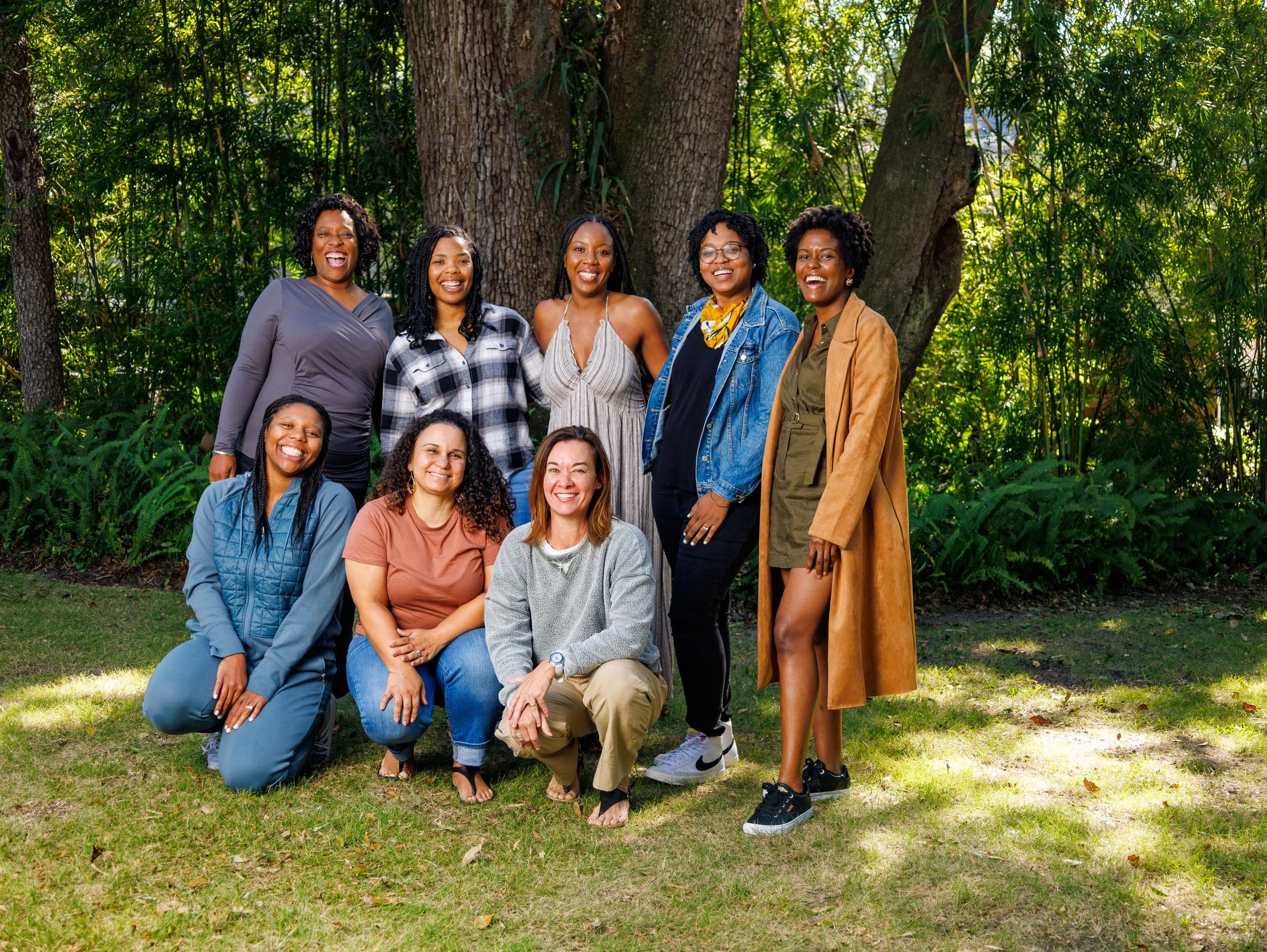 Group of ten women smiling and posing outdoors in a park with trees and greenery.