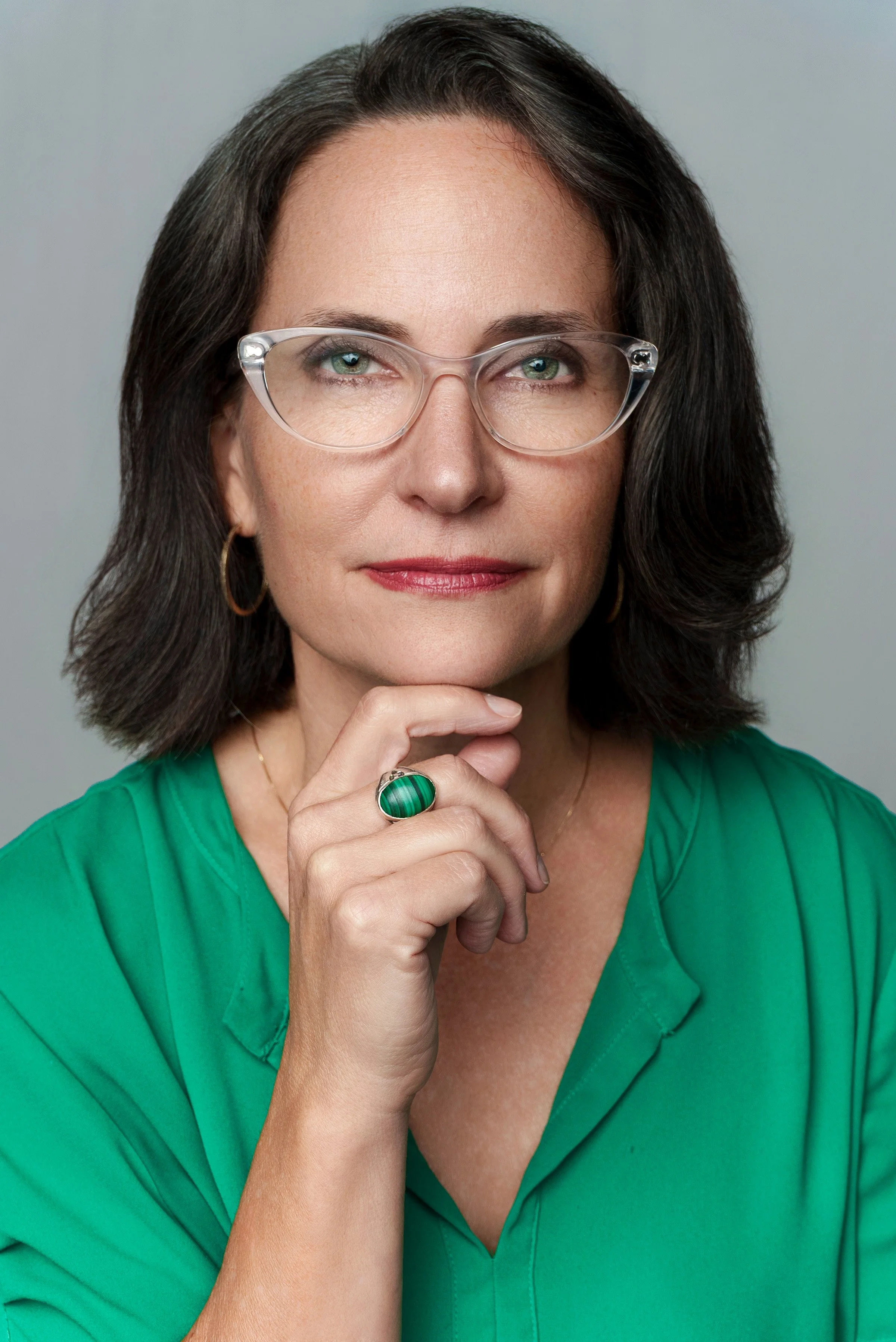 A woman with dark hair, wearing glasses and a green blouse, poses with her hand near her chin, showing a large green ring.