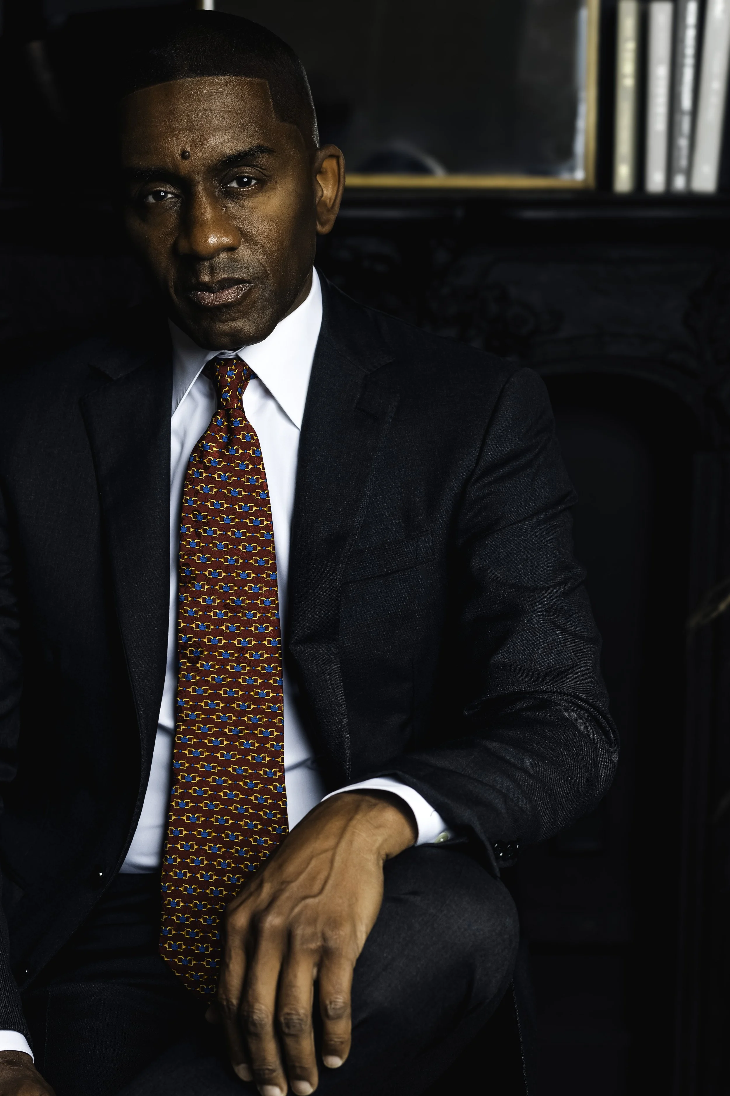 A man in a dark suit sitting in front of a mirror with bookshelves in the background.