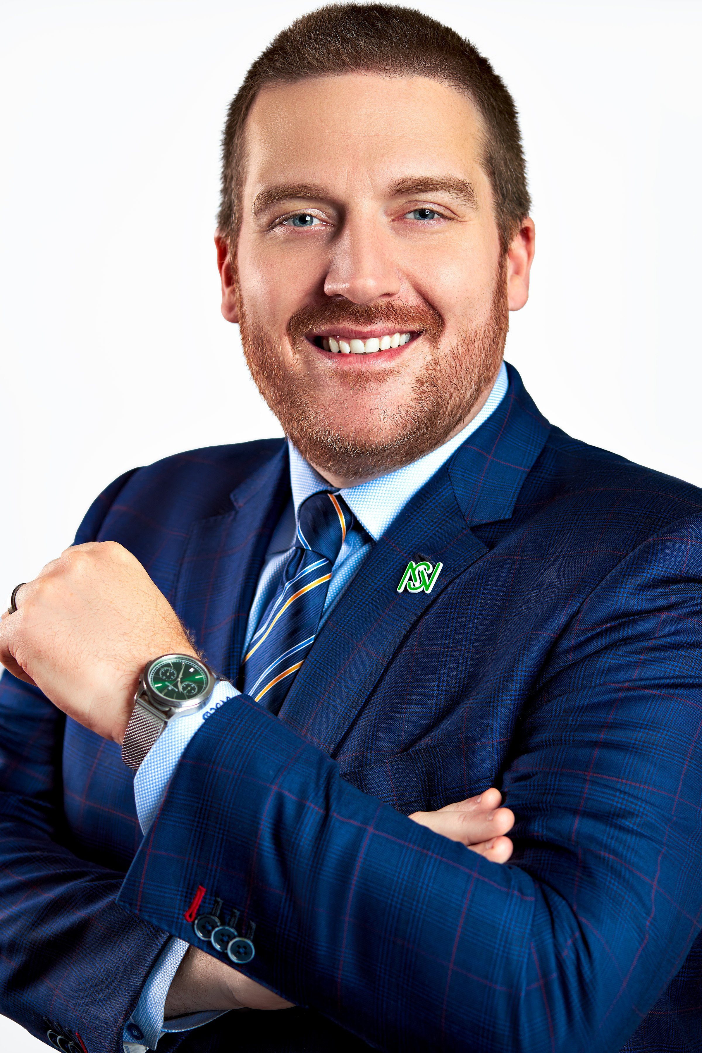 A man in a blue checked suit and tie, smiling with arms crossed, wearing a silver watch, and a lapel pin with the North Carolina State University logo.