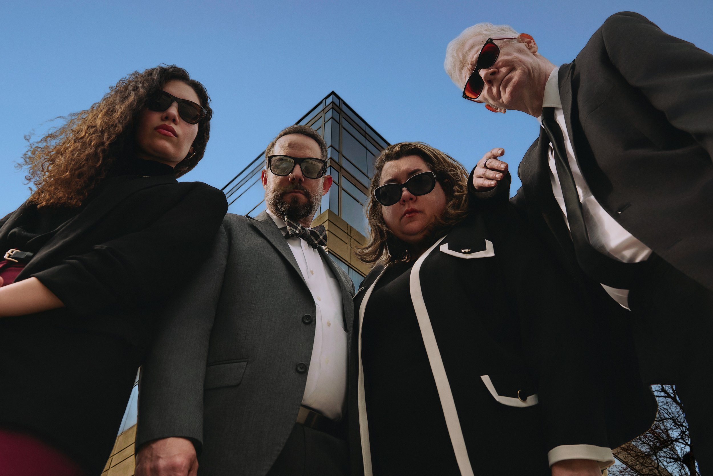A group of four people looking down at the camera, wearing sunglasses, with a modern building and blue sky in the background.