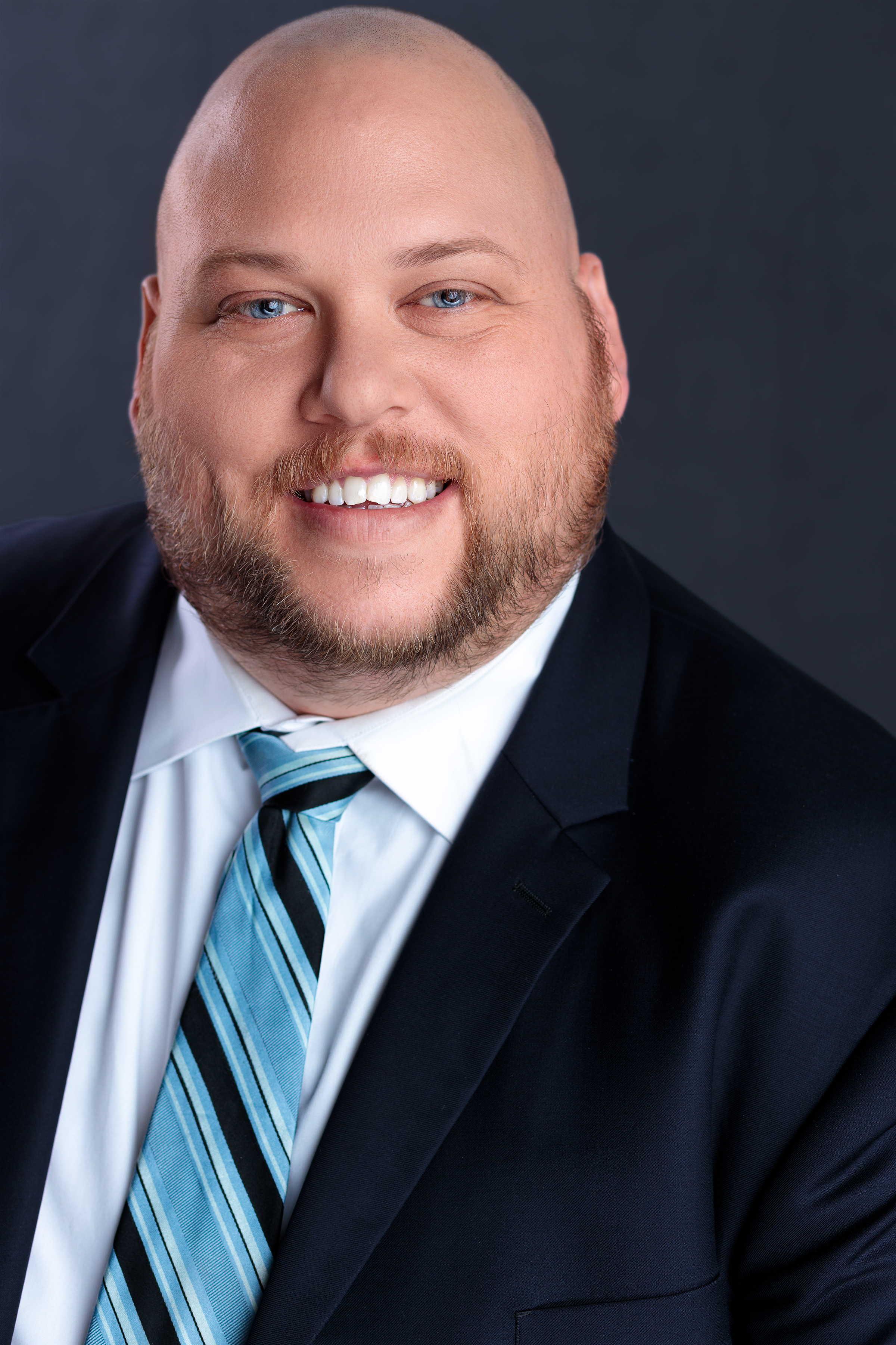 Close-up professional headshot of a man with a bald head and a beard, smiling, wearing a dark suit, white shirt, and striped blue tie, against a dark background.