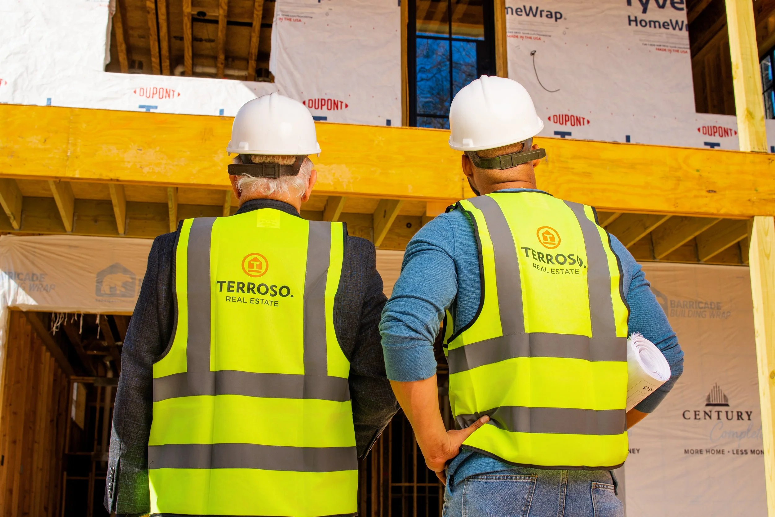 Two construction workers wearing yellow safety vests and white hard hats standing in front of a building under construction.