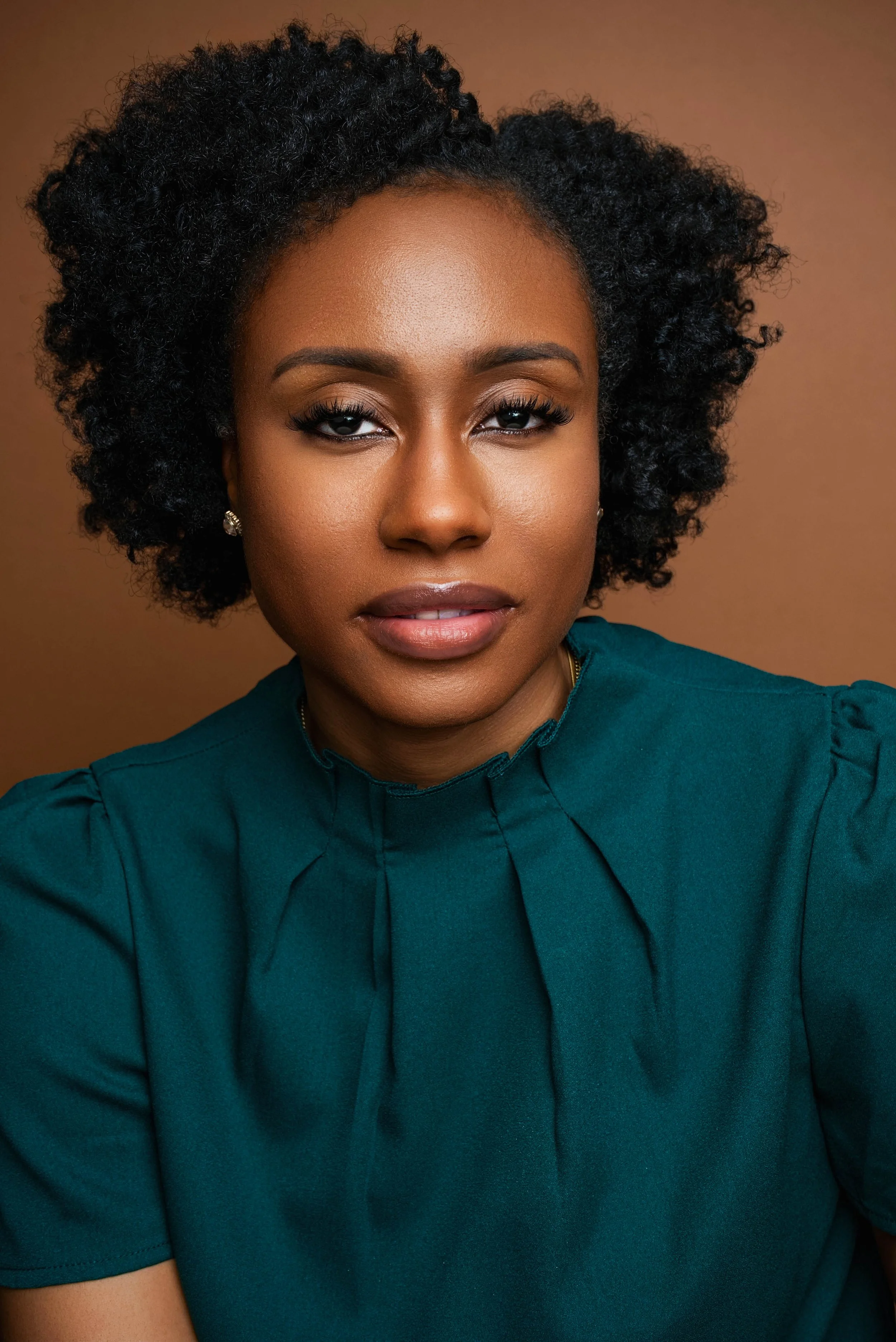 A woman with dark, curly hair styled to one side, wearing a teal blouse with pleated details and small earrings, looking directly at the camera with a confident expression.