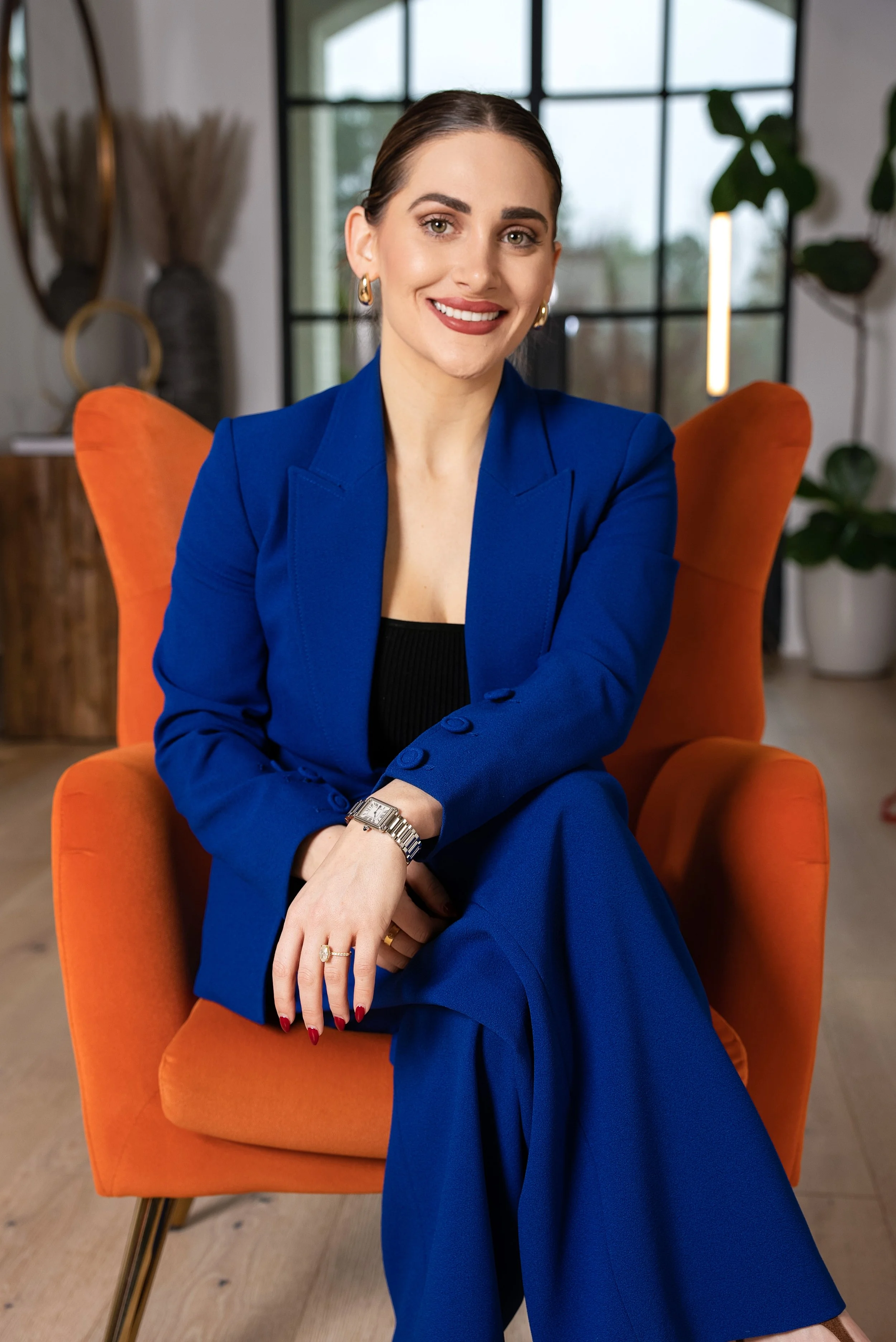 A woman with dark hair pulled back, wearing a royal blue suit, sitting on an orange chair in a modern room with large windows and potted plants, smiling at the camera.