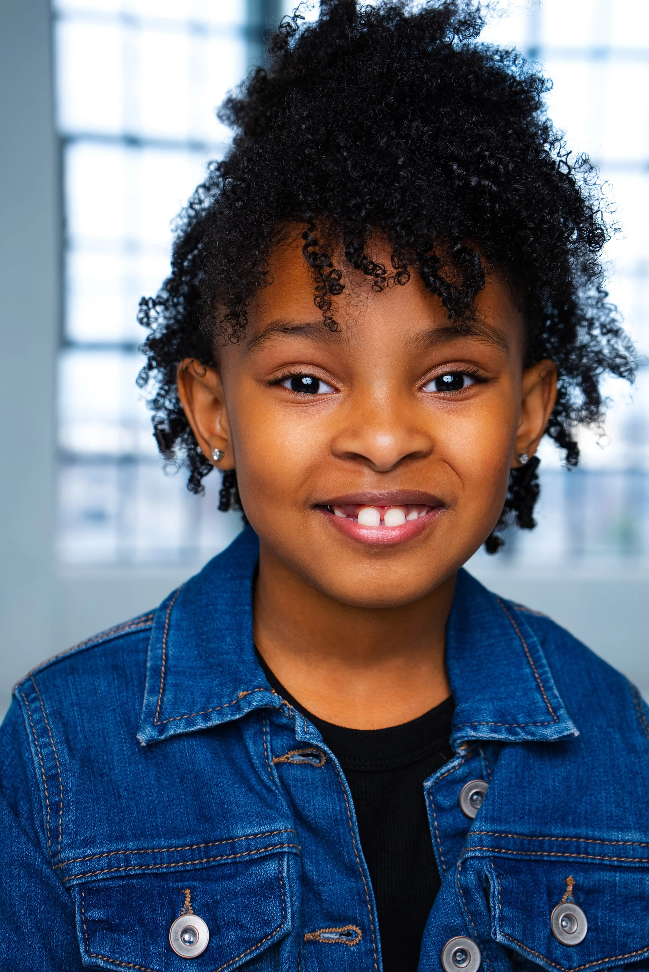 A young girl with curly black hair smiling, wearing a blue denim jacket and a black shirt, indoors with a blurred window in the background.