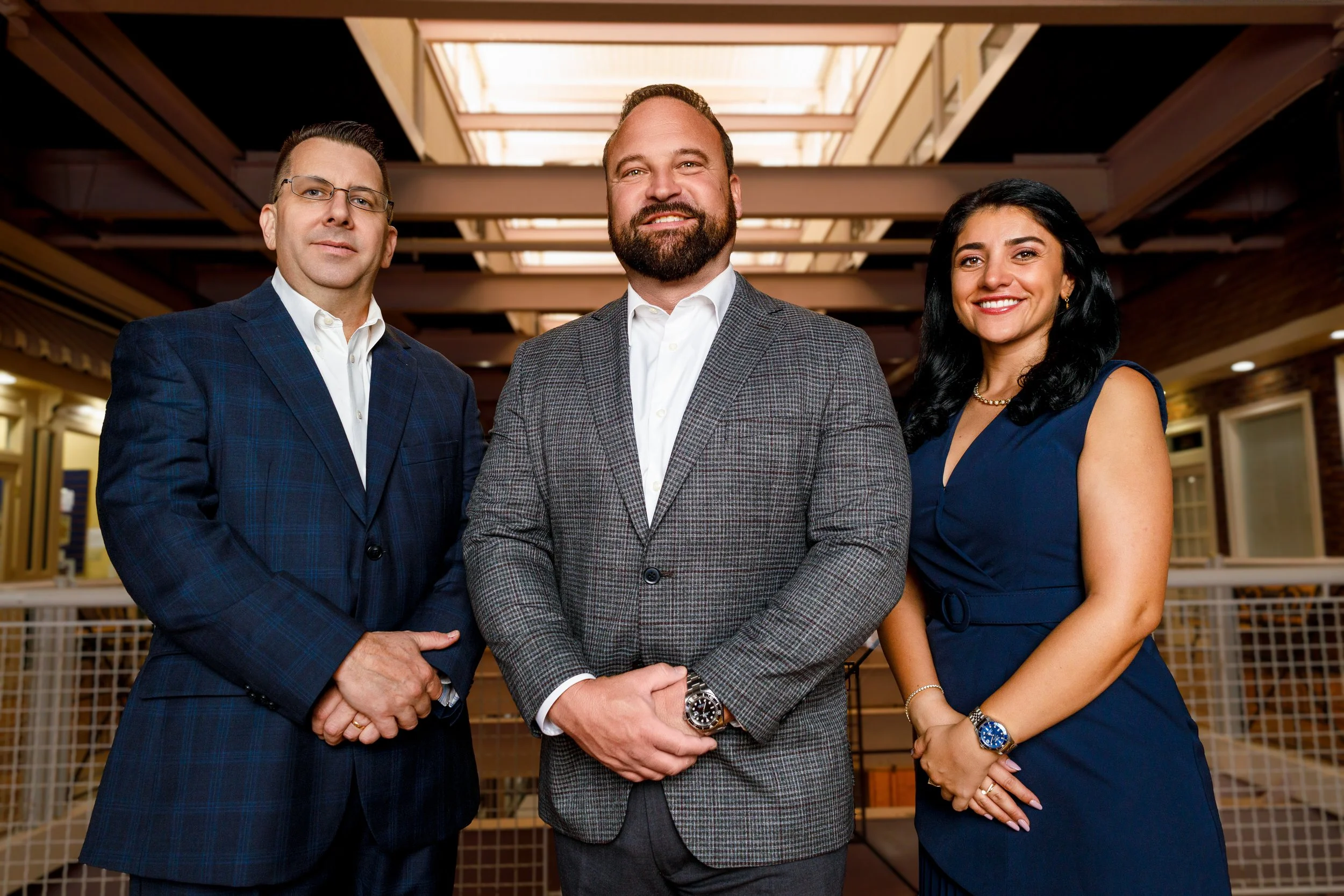 Three professionally dressed people, two men and one woman, standing together indoors with open ceiling and beams in the background.