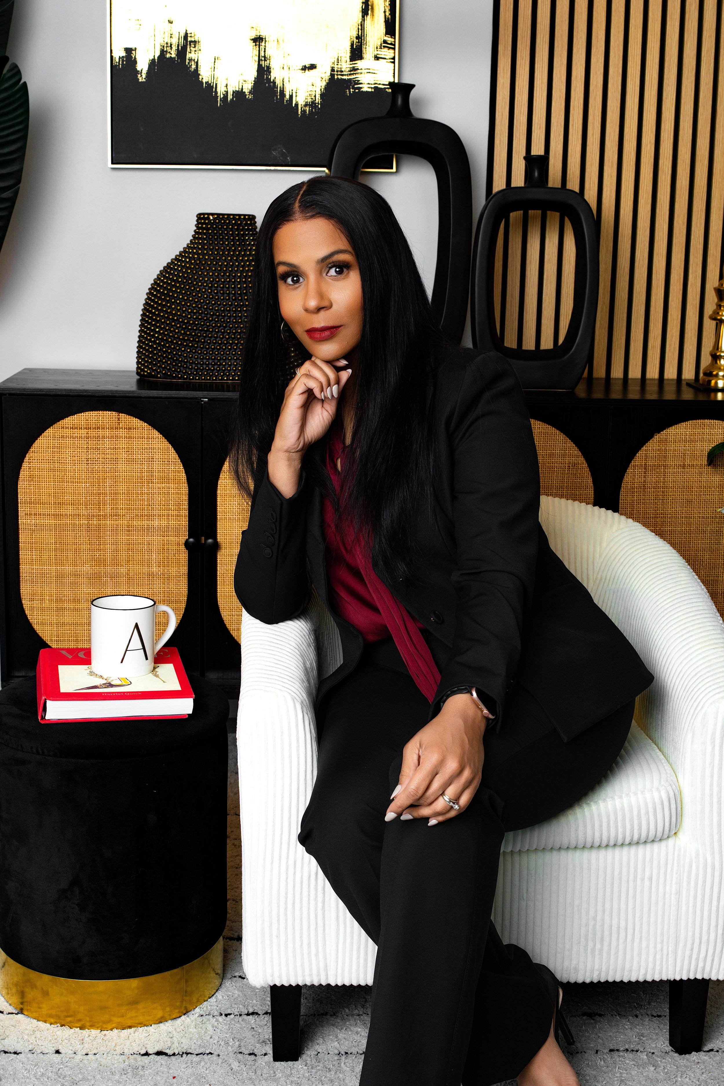 A woman sitting on a white sofa in a modern, stylish room with decorative vases and abstract artwork on the wall. She is wearing a black blazer and dark pants, with a maroon top, and has long black hair. There is a black side table with a white mug, 