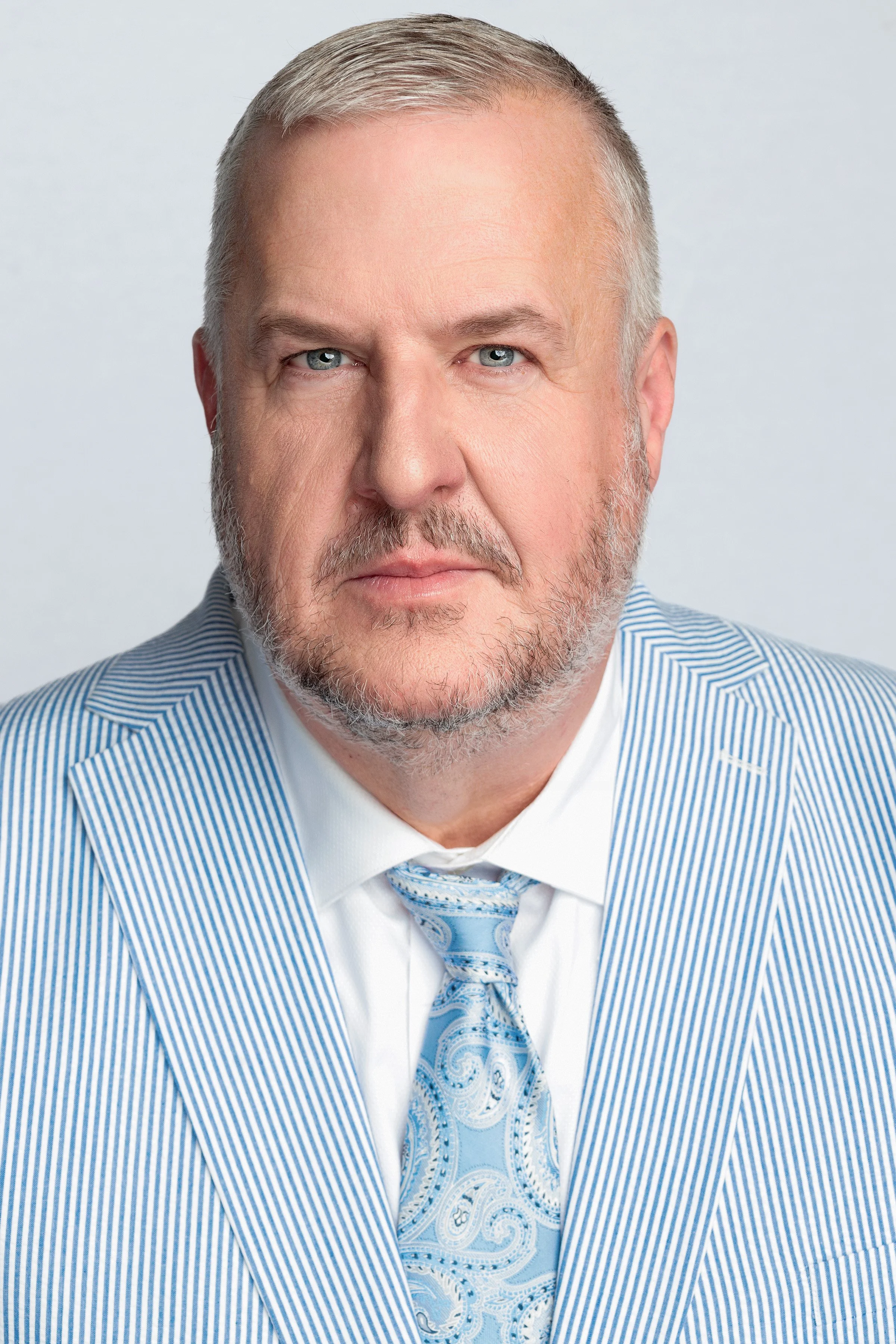 A middle-aged man with gray hair and beard, wearing a light blue and white pinstripe suit with a matching tie, posing against a light gray background.