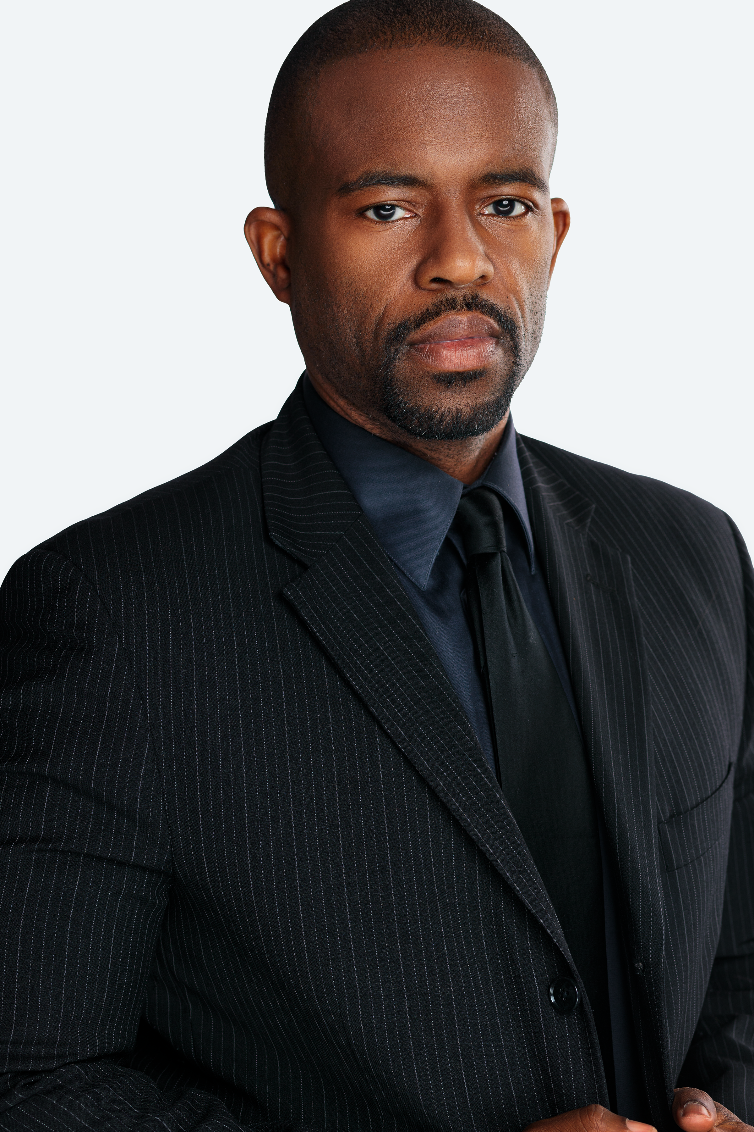 A serious-looking African American man in a dark pinstripe suit, black shirt, and black tie against a plain light background.