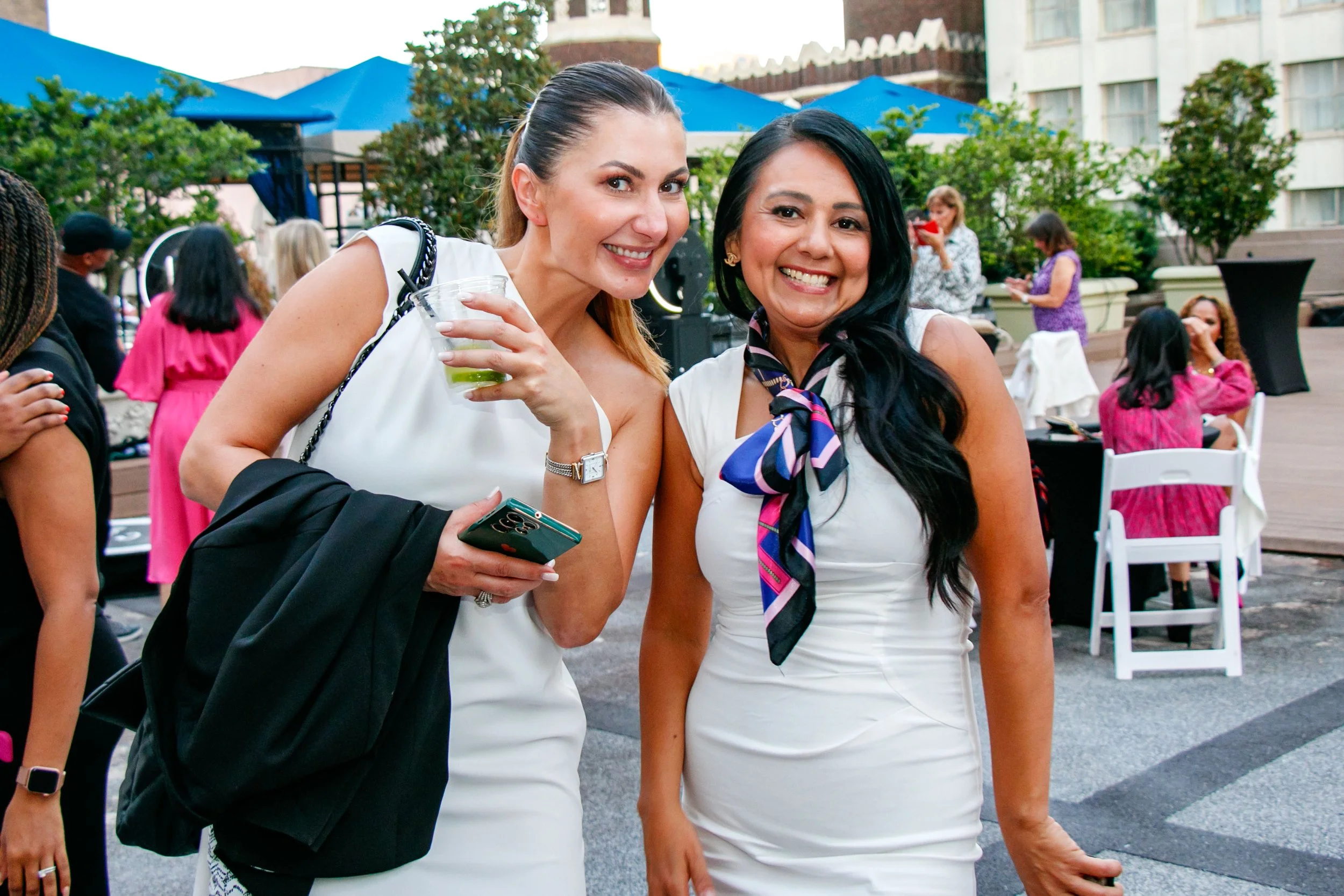 Two women smiling at an outdoor event, one holding a drink and a phone, with several people and tables in the background.